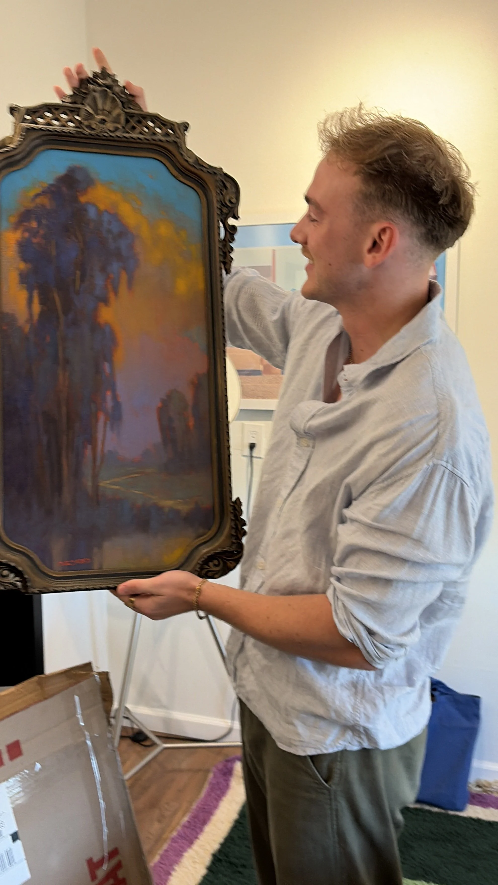 A man with light brown hair holding a framed landscape painting. The painting depicts trees with a colorful sky.