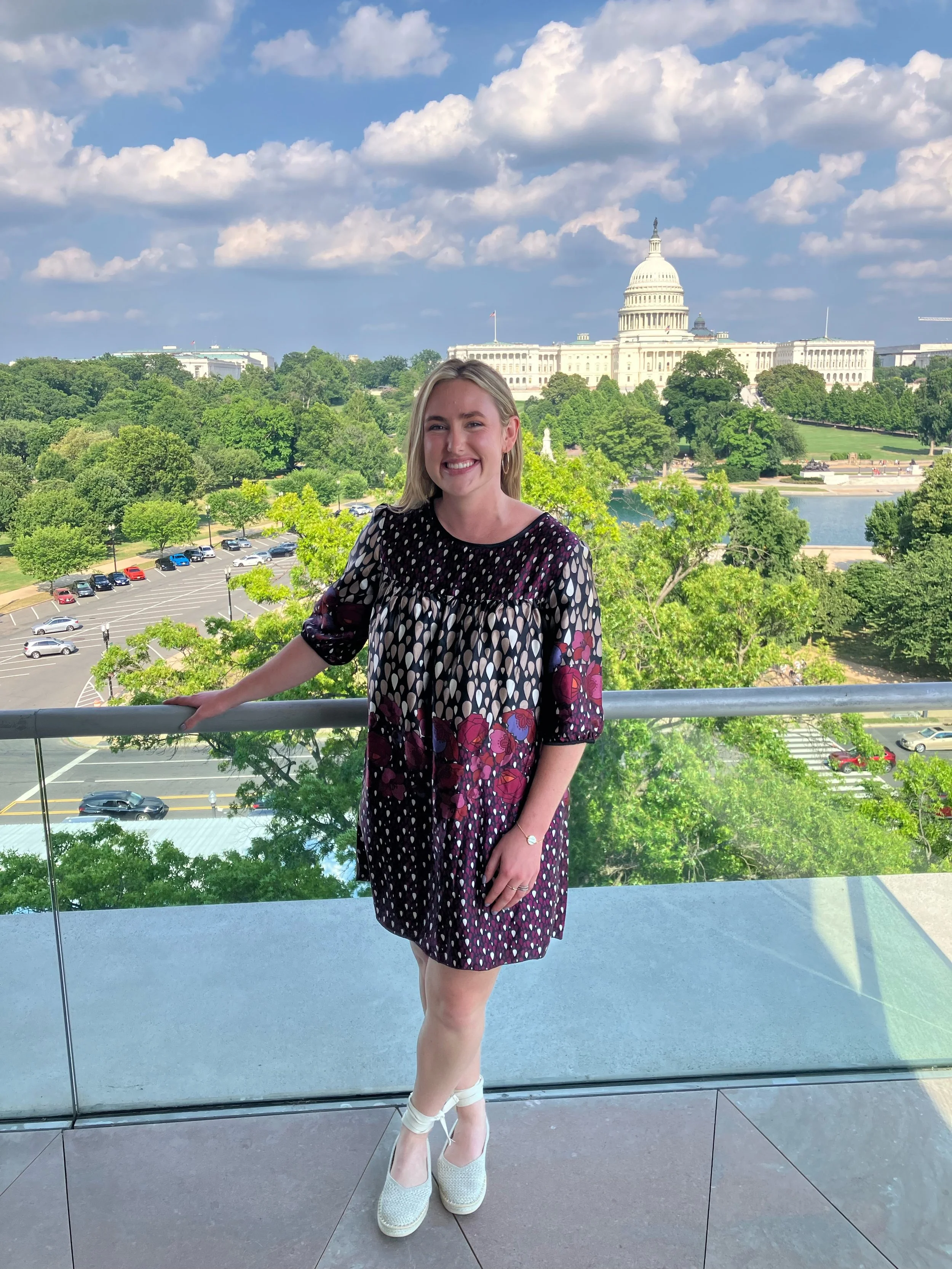 A young woman with blonde hair smiling while standing on a balcony overlooking the U.S. Capitol building in Washington, D.C., with trees and a parking lot below on a sunny day with partly cloudy skies.