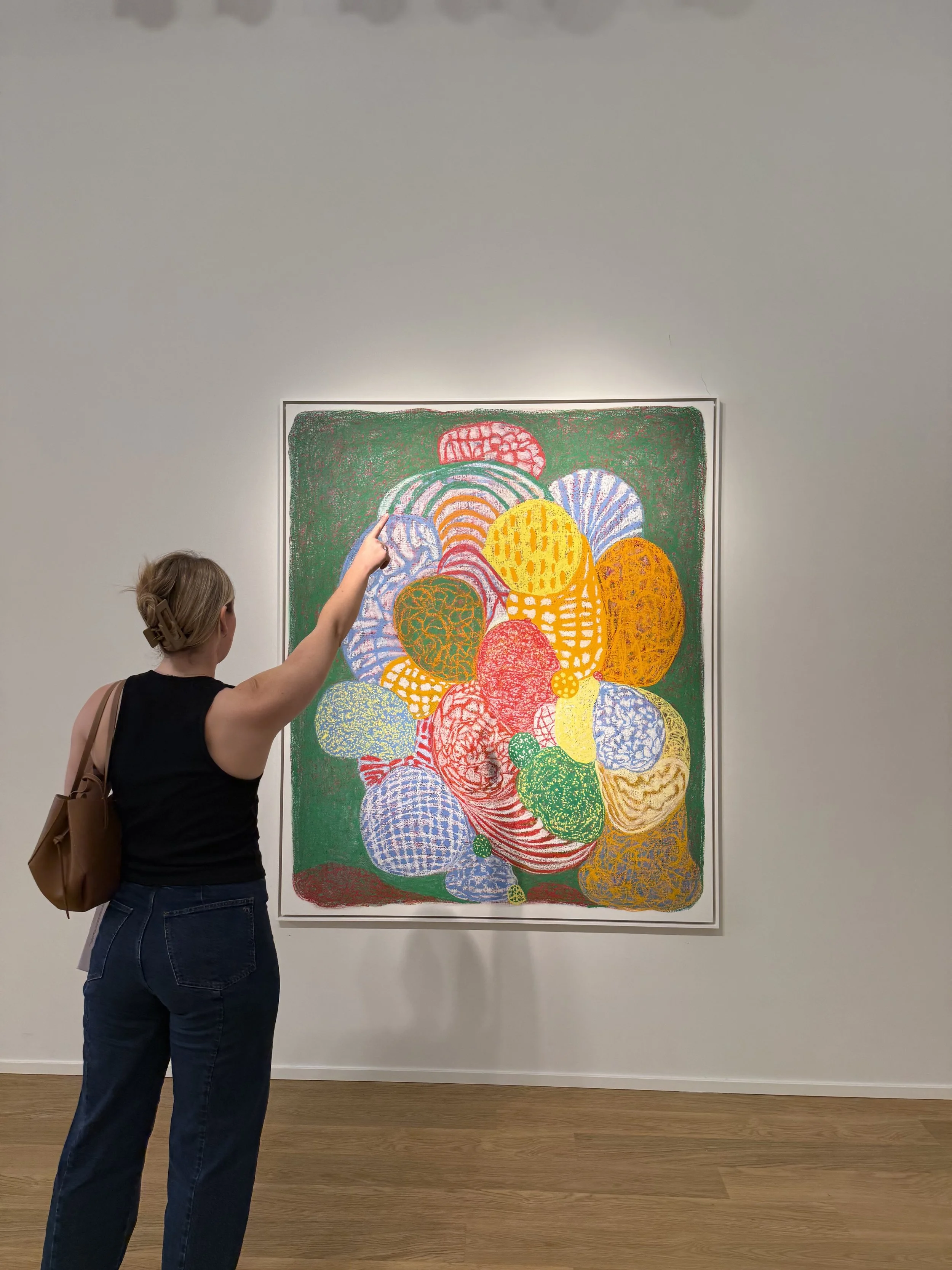 A woman standing in front of a colorful abstract painting in a gallery, pointing at the artwork with her right hand, wearing a black sleeveless top, jeans, and carrying a brown shoulder bag.