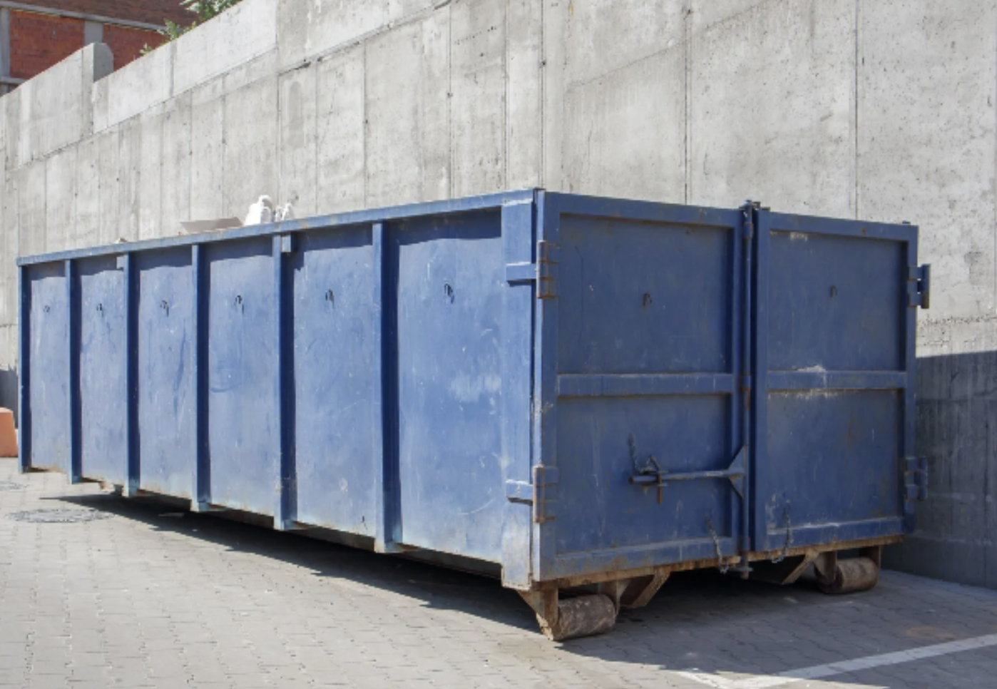 A large blue industrial dumpster on wheels next to a concrete wall.