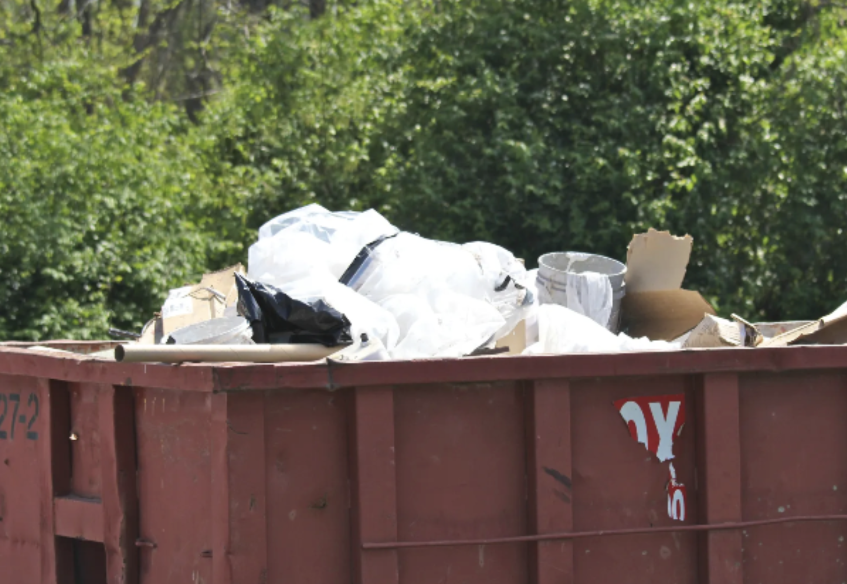 A red dumpster filled with trash, including plastic bags, paper, and other debris, set against a background of green trees.