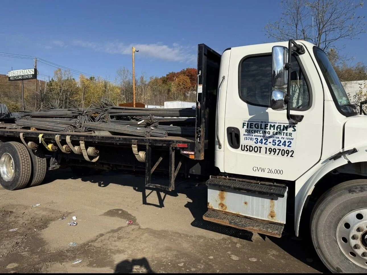 White flatbed truck with black bed carrying metal rods, parked outdoors at a recycling center, with trees and a blue sky in the background.