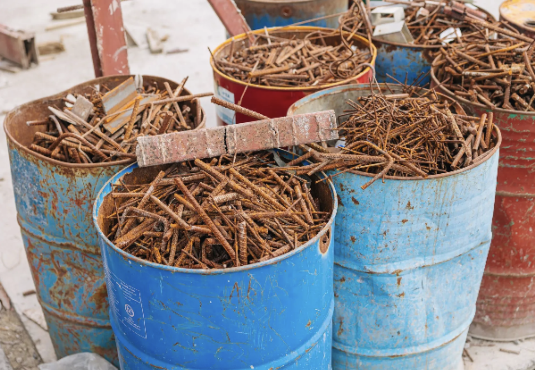 Five blue and red metal barrels filled with rusty steel rebar scrap, placed outdoors on a construction site.