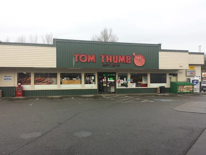 Exterior of a grocery store named 'Tom Thumb' with a green and beige facade, red signage, and a small grocery cart area outside.