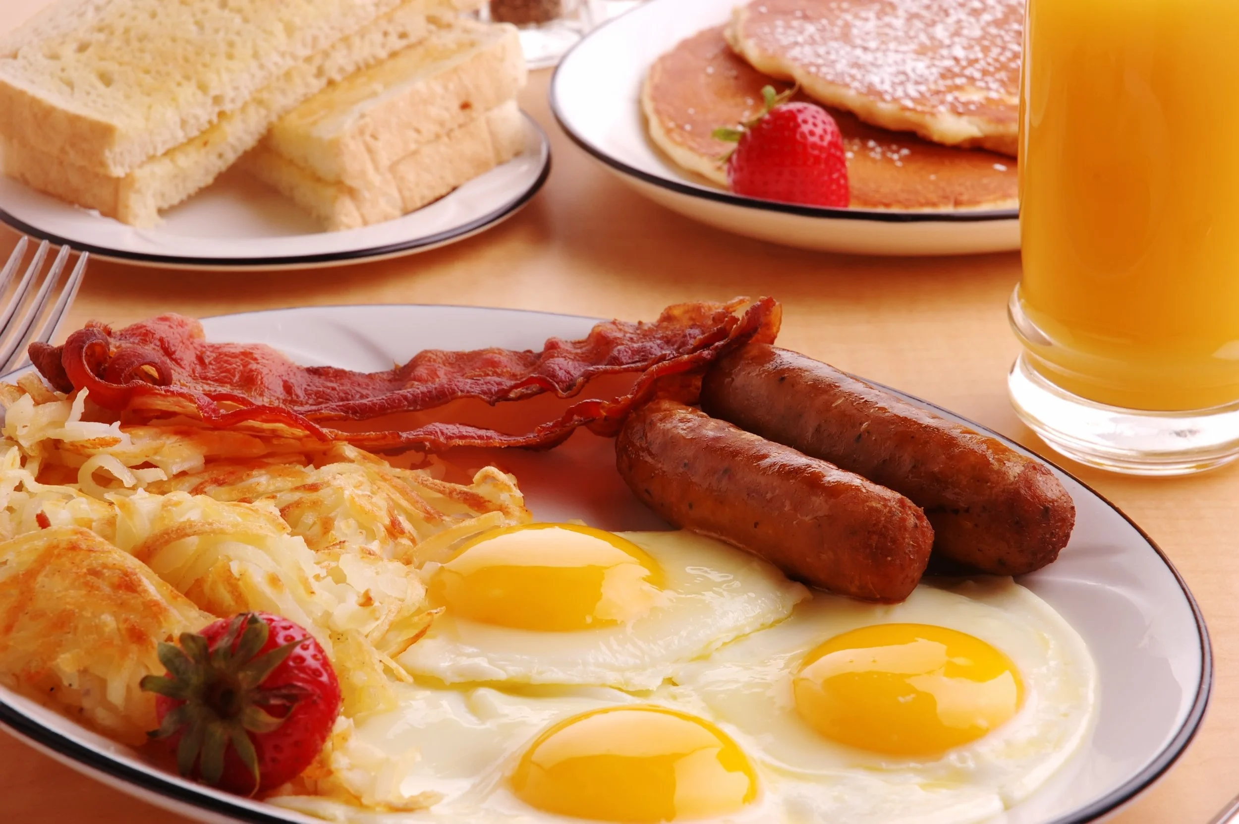 A breakfast plate with sunny side up eggs, bacon, sausages, hash browns, and shredded cheese, accompanied by a glass of orange juice, with additional plates of pancakes, toast, and strawberries in the background.