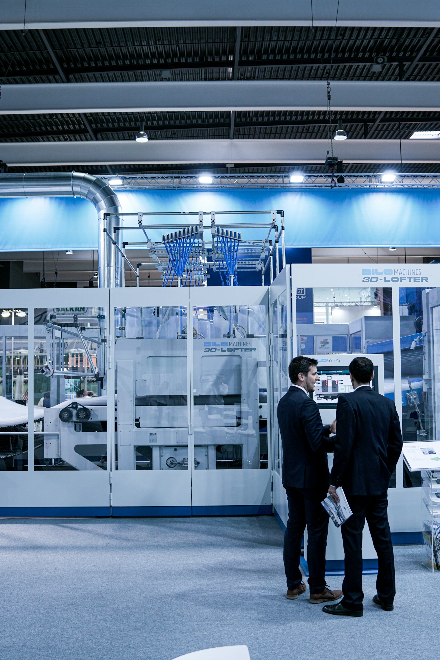 Two men in business suits are conversing and exchanging information at a trade show or exhibition booth that showcases industrial machinery, including a large white 3D-LATOR machine and other equipment.