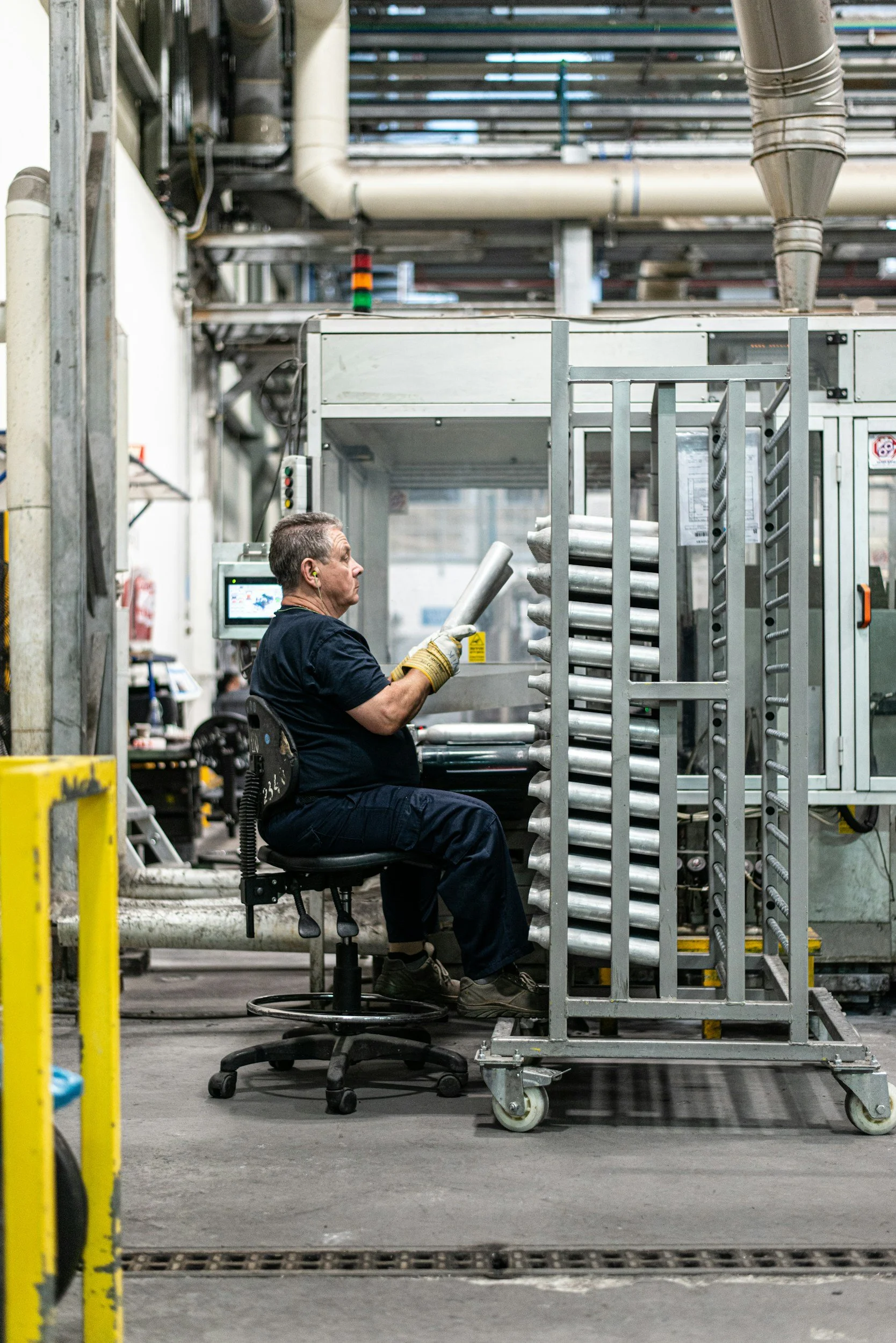 A worker in black uniform and ear protection seated on a rolling chair, working with metal pipes in an industrial setting with ductwork and machinery overhead.