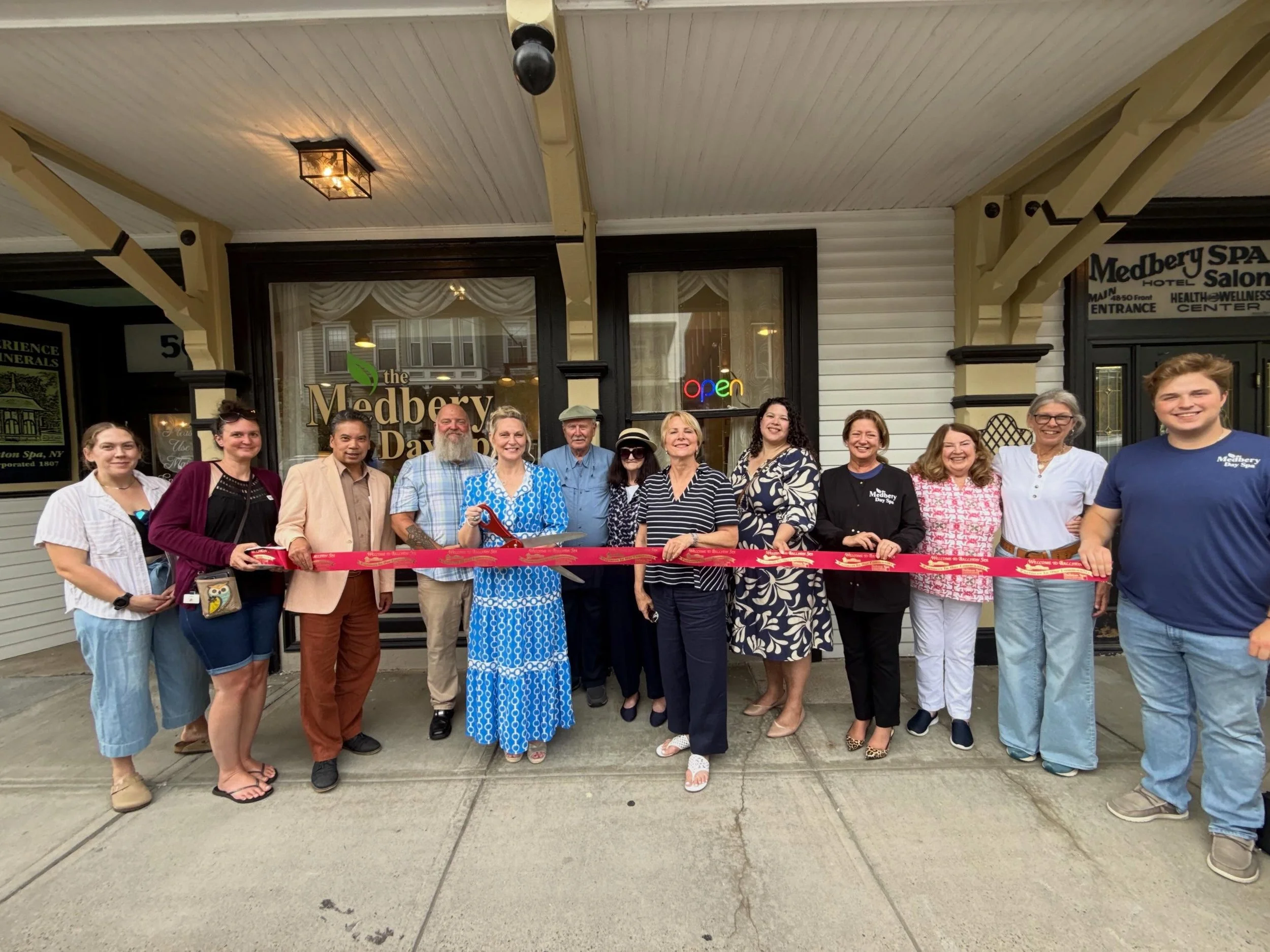 Group of people standing in front of the Medbery Day Spa, holding a red ribbon for a ribbon-cutting ceremony.