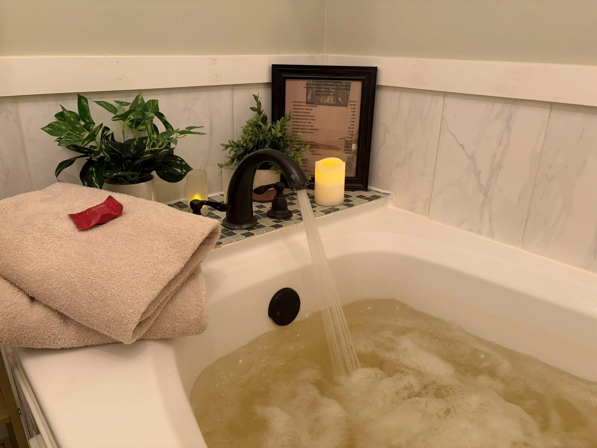 A bathtub filled with water with a headrest on the side, candles around it, a small wooden tray with a glass jar, a candle, and some snacks. There is a sign on a stand next to the bathtub, and a black chair partially visible.