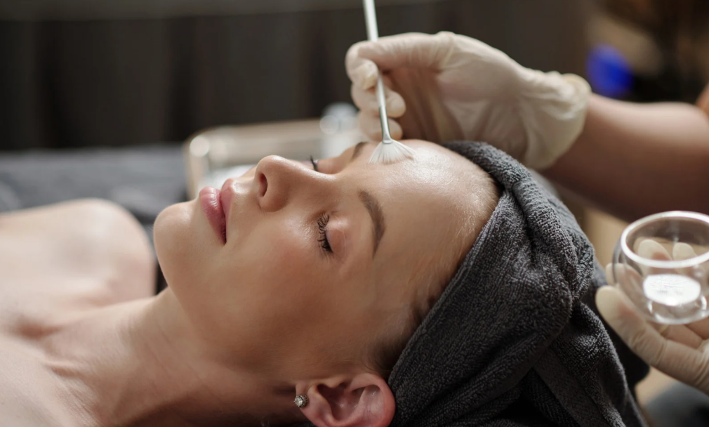 A woman receiving a facial treatment at a spa or salon, with a towel or headband on her head, eyes closed, and a beauty professional using a brush to apply a skincare product.