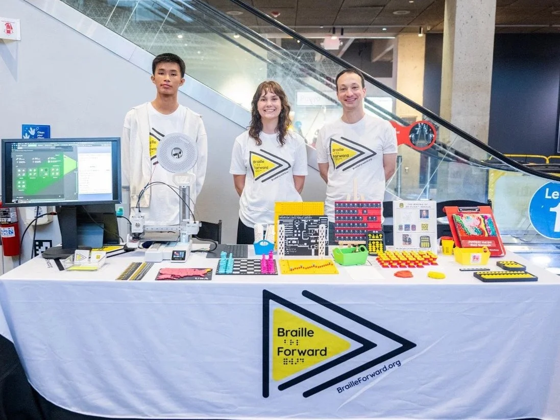 Richard He, Coco Fraser, and Alex Eisenberg stand behind a table with Braille Forward products and a 3D-printing demonstration at the Museum of Science's Disability Pride 2026. Photo © Ashley McCabe