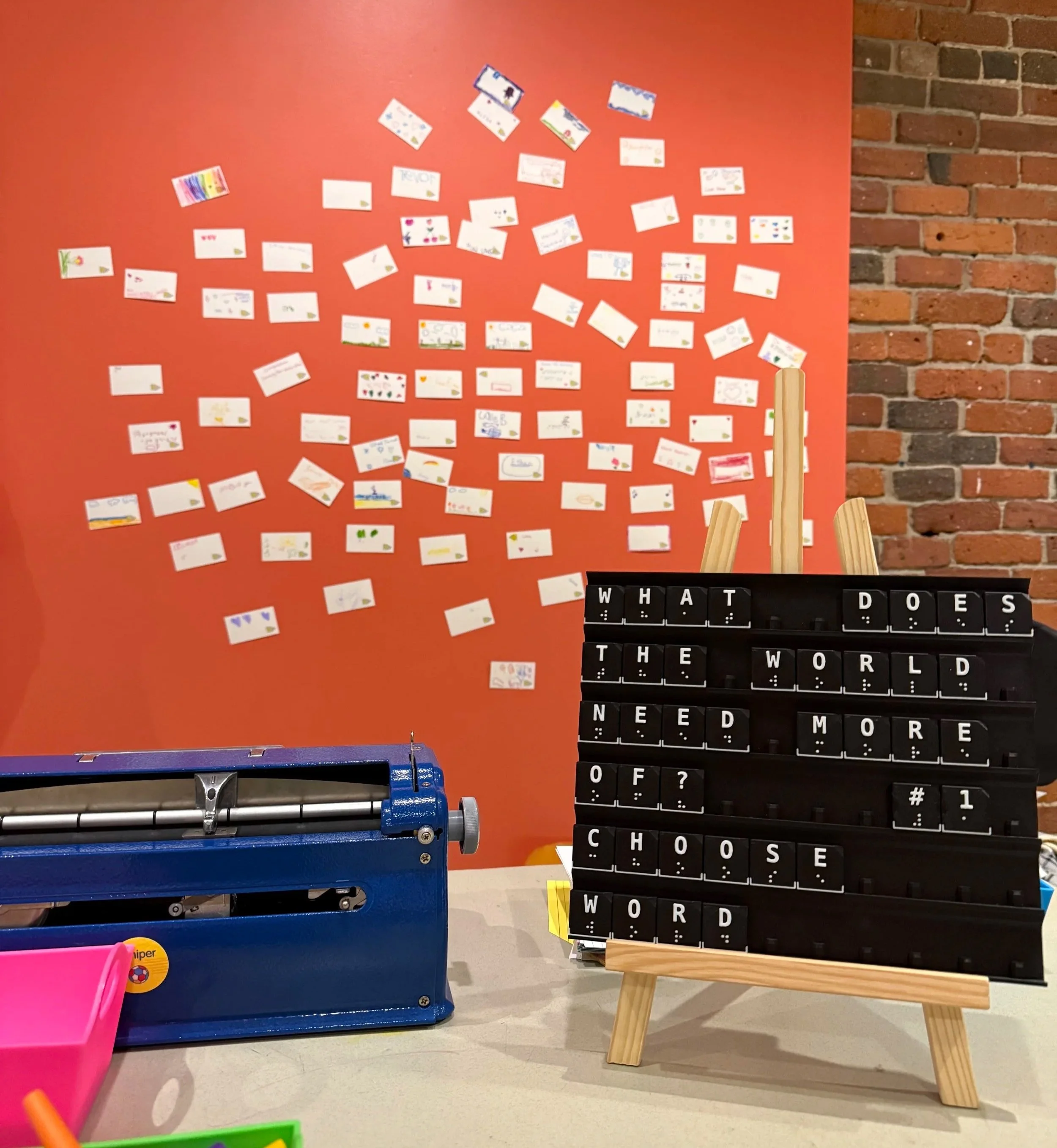 A sign (made with Braille Forward's Letter Farm frame and tiles) asks "what does the world need more of?". Behind the sign, there is a wall of notecards with different answers to this question, made with the Perkins Brailler.