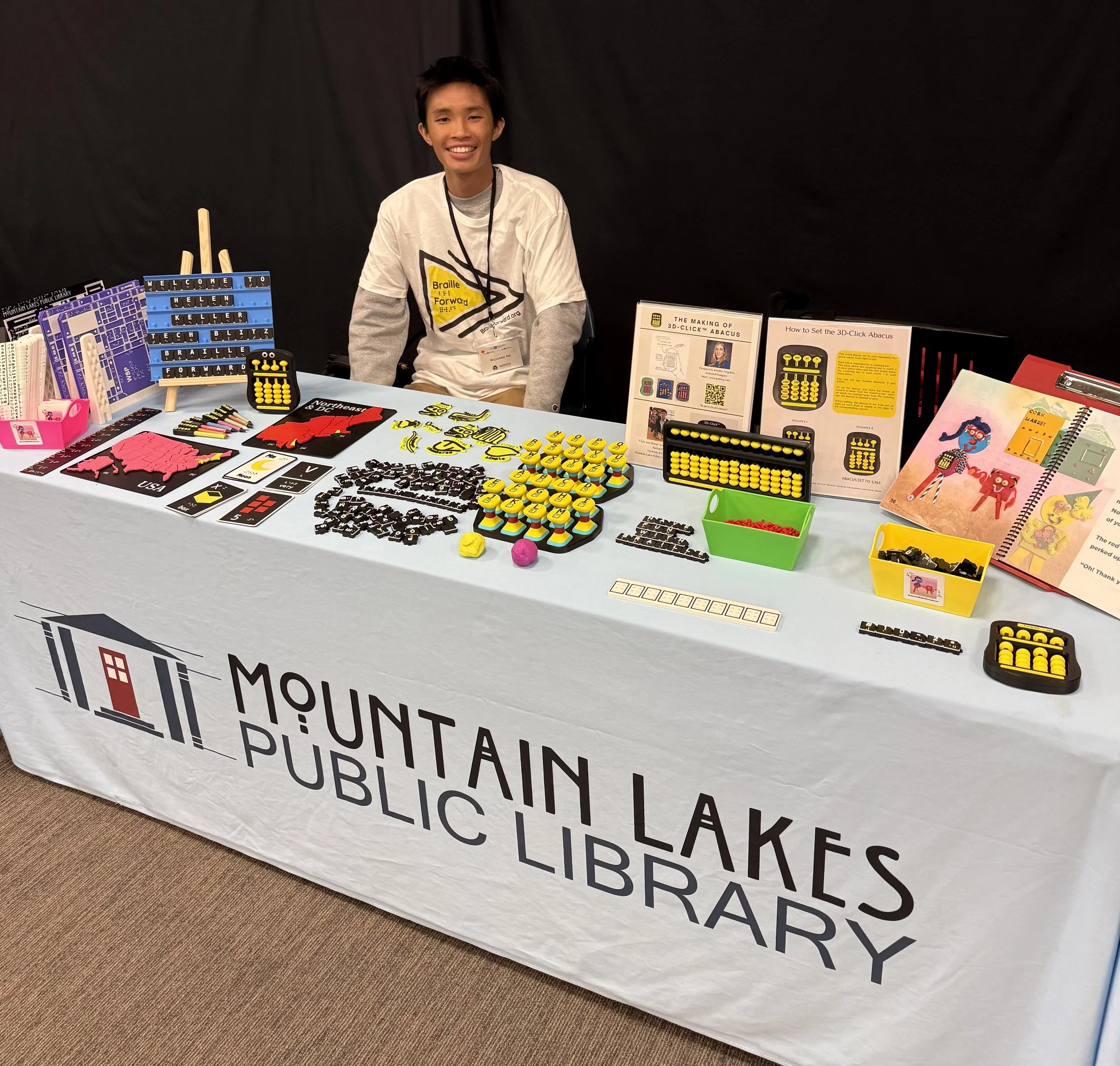 Richard He sitting behind a table with a variety of Braille Forward's tactile learning materials.