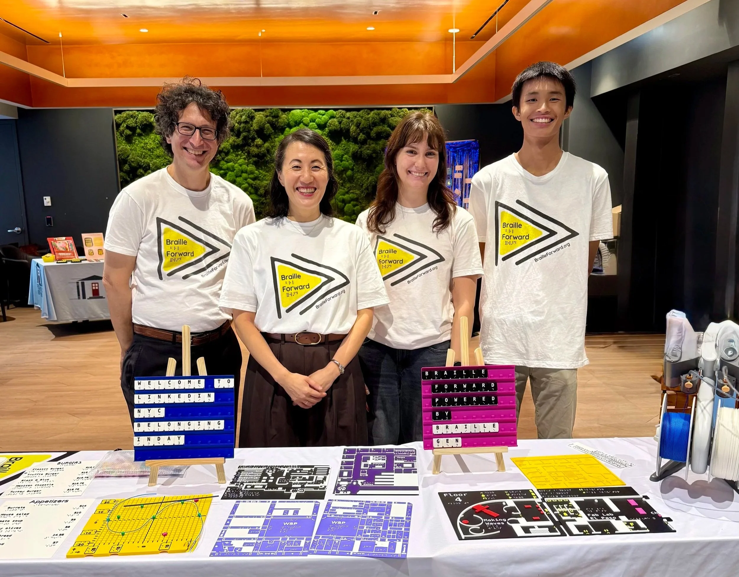 The Braille Forward team standing in front of a variety of tactile maps and other universally designed resources. From left to right: Ian Matty, Chi-Hoon Kim, Coco Fraser, and Richard He.