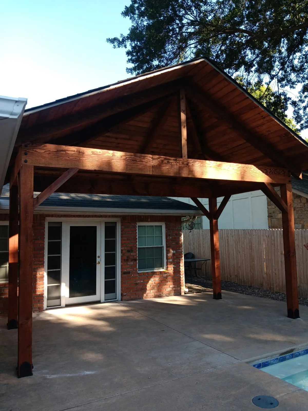 Backyard patio with a new wooden pergola over the concrete slab, brick house wall, sliding glass door, and a fenced yard with trees.