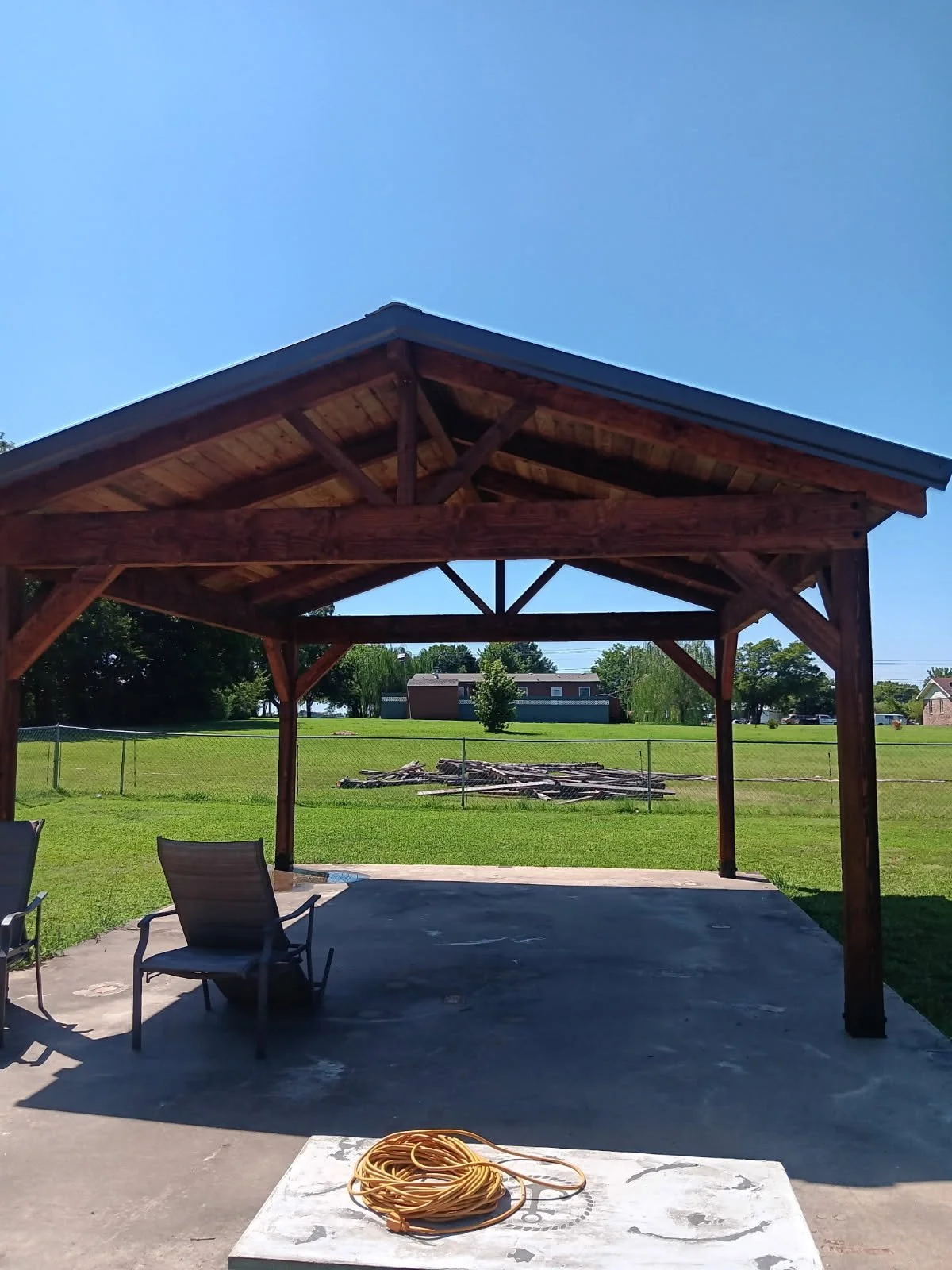 A wooden gazebo with a metal roof in a backyard, with chairs on the concrete patio, a grassy yard, trees, and a house in the background under a clear blue sky, and a yellow extension cord on a white table in the foreground.