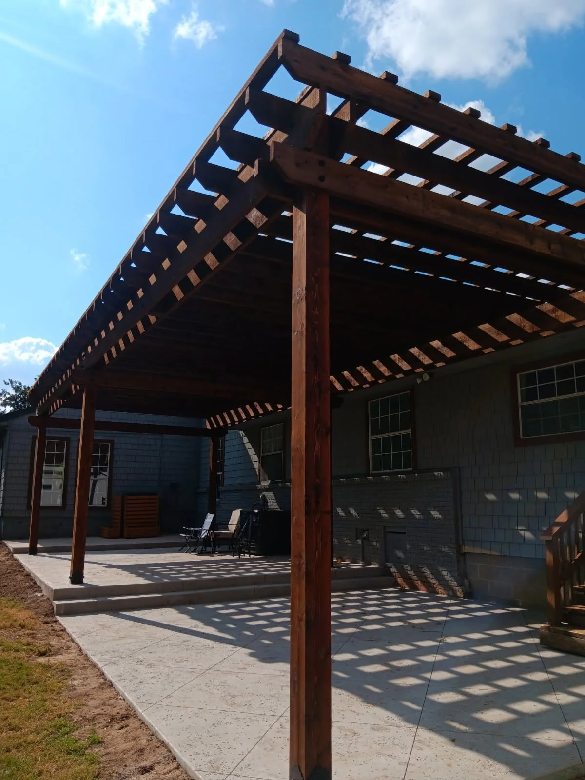 A backyard patio with a new wooden pergola, outdoor chairs, and a table. The house is visible in the background with windows and gray siding. The sky is blue with some clouds.