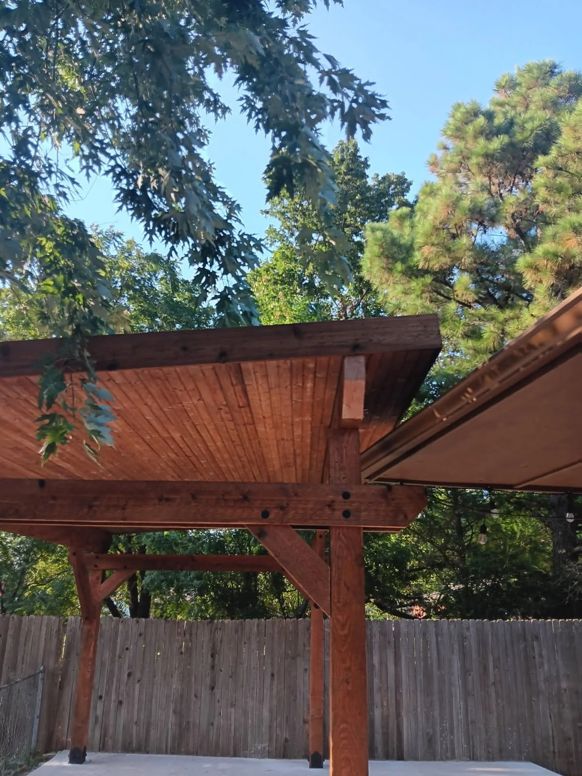 Wooden deck or pergola structure with a roof, surrounded by green trees and a wooden fence in the background, under a clear blue sky.