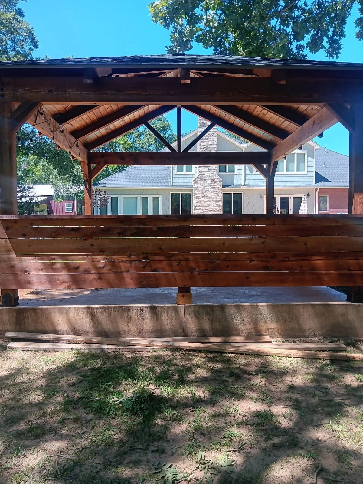 View through a wooden gazebo with a built-in bench to a suburban backyard with a house featuring a stone chimney and grey siding, surrounded by green trees and a clear blue sky.