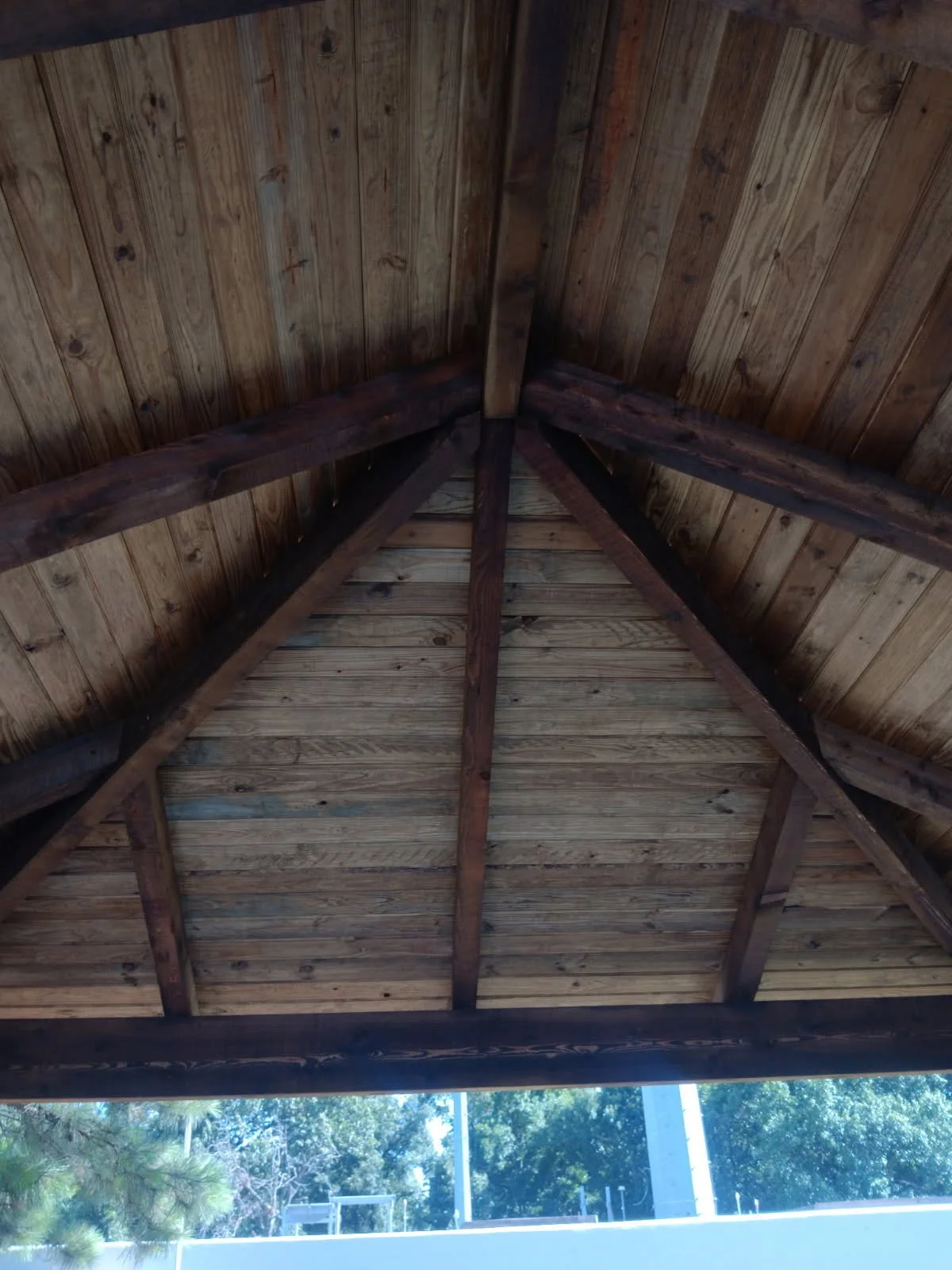 View of a wooden pavilion ceiling with exposed beams and planks, with greenery and a parking lot visible outside.