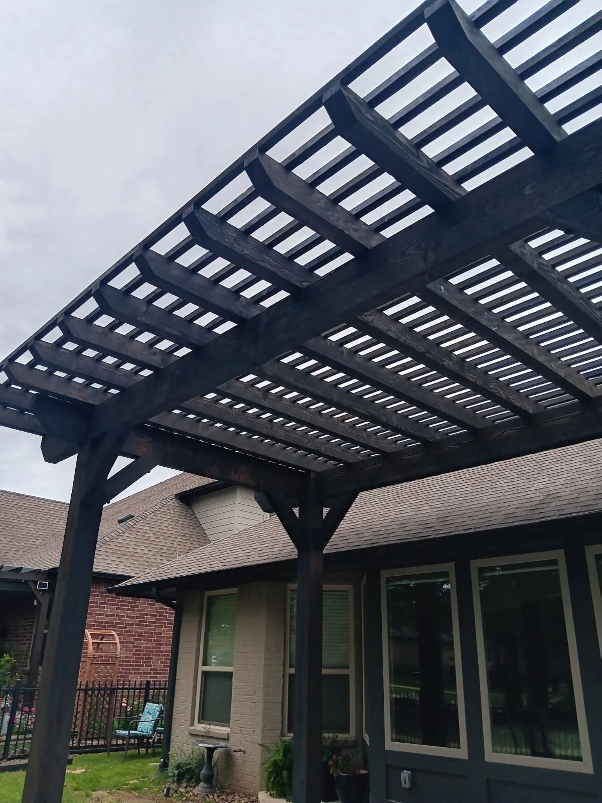 Wooden outdoor pergola over patio with house, window, brick wall, and garden furniture visible.