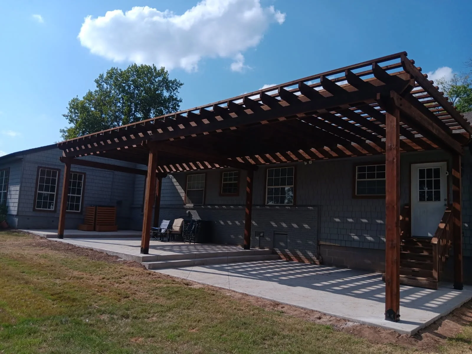 Back of a house with a wooden pergola shading a patio area with outdoor furniture, three steps leading down to a grassy yard, and a blue sky with clouds.
