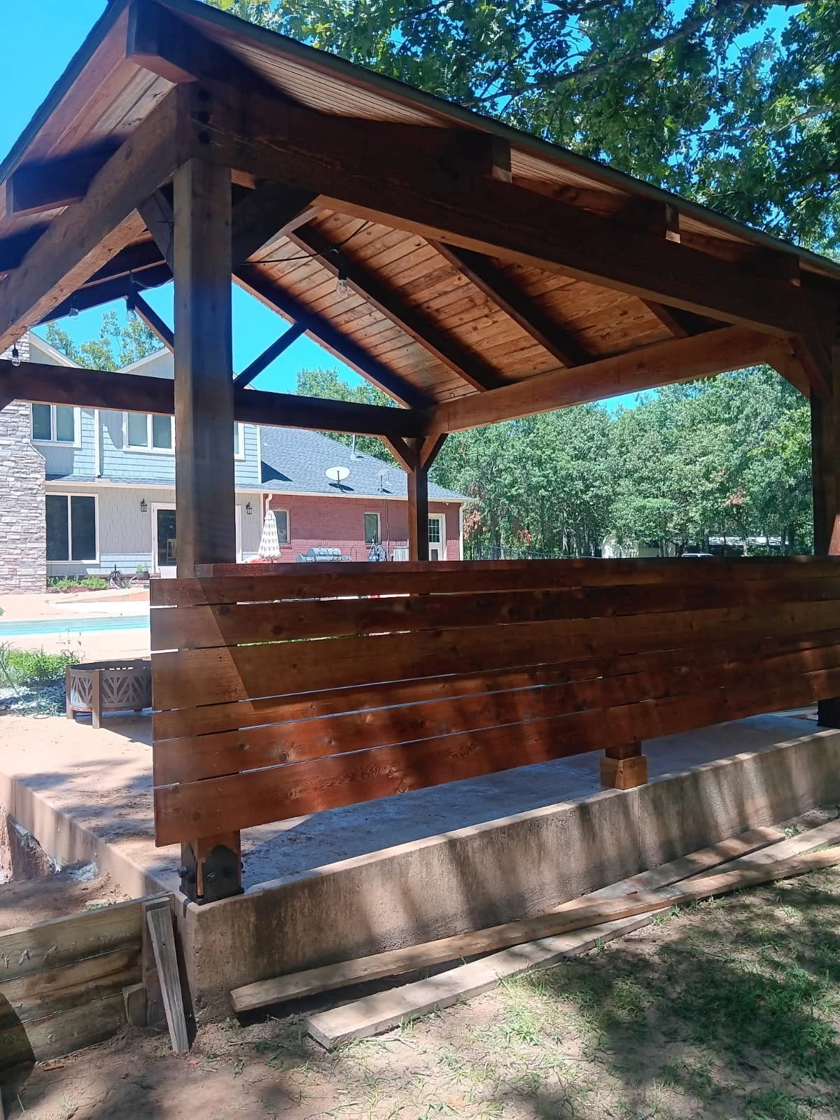 Wooden gazebo with a sloped roof located in a backyard with houses and trees in the background.