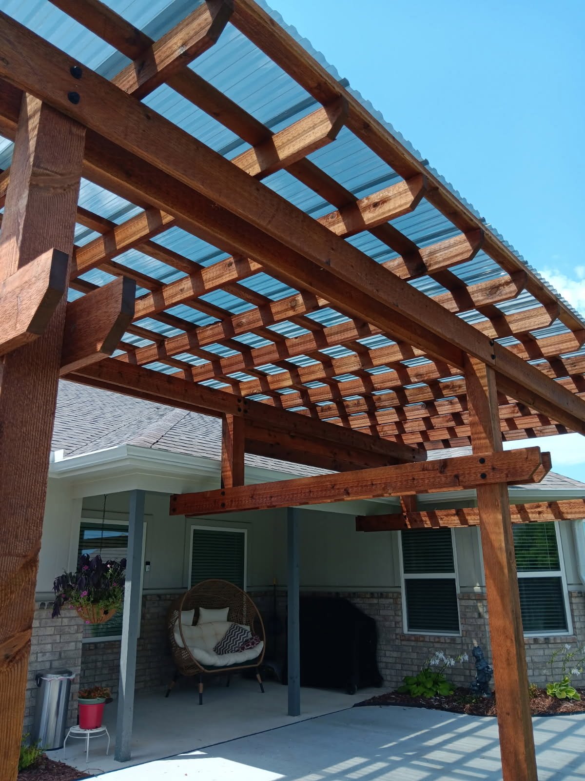 Wooden pergola with a plastic roof over a backyard patio area with outdoor furniture and plants.
