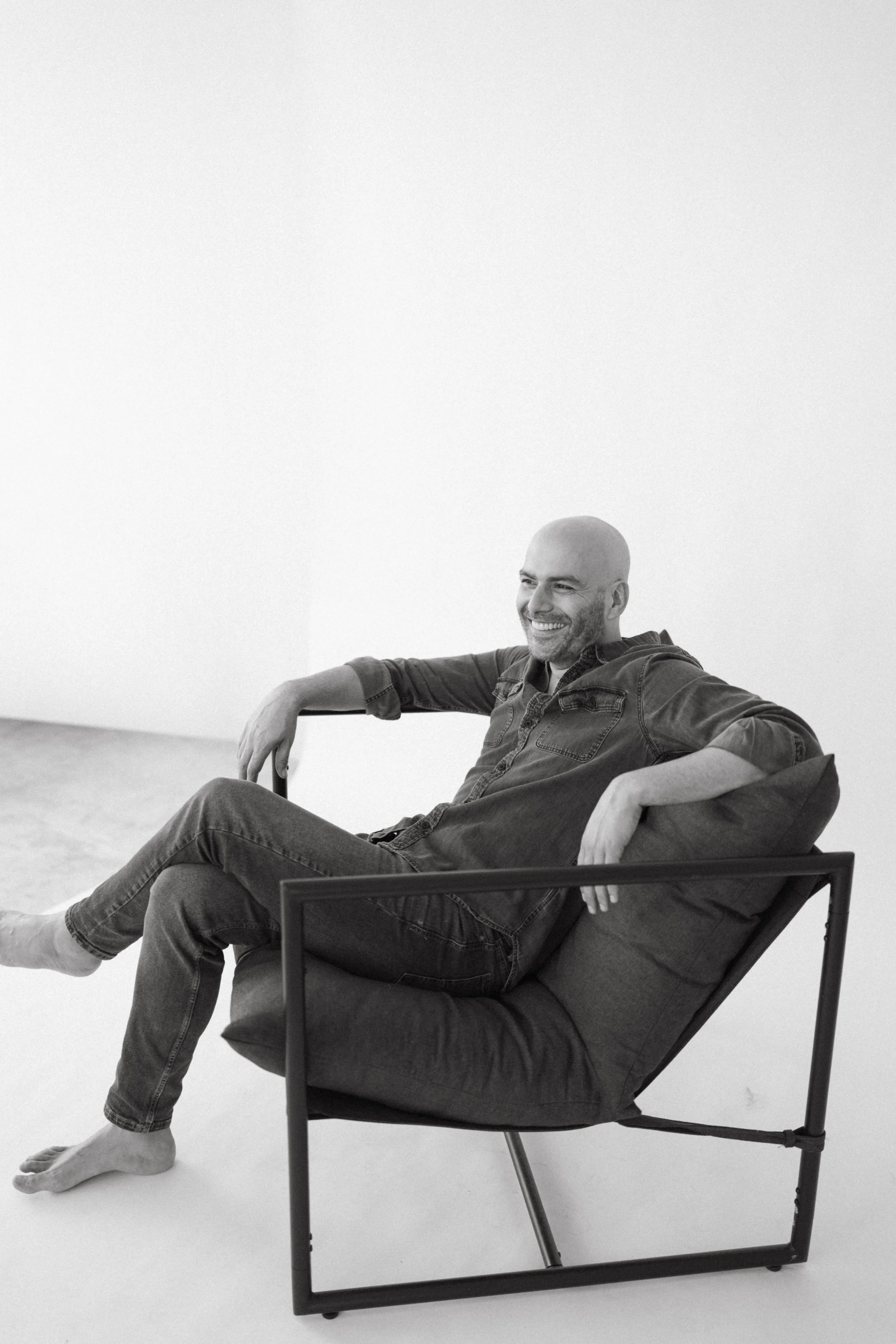 A man with a shaved head and a beard sitting in a modern chair, smiling and relaxing with his arms resting on the armrests, in a minimalist room with white walls.