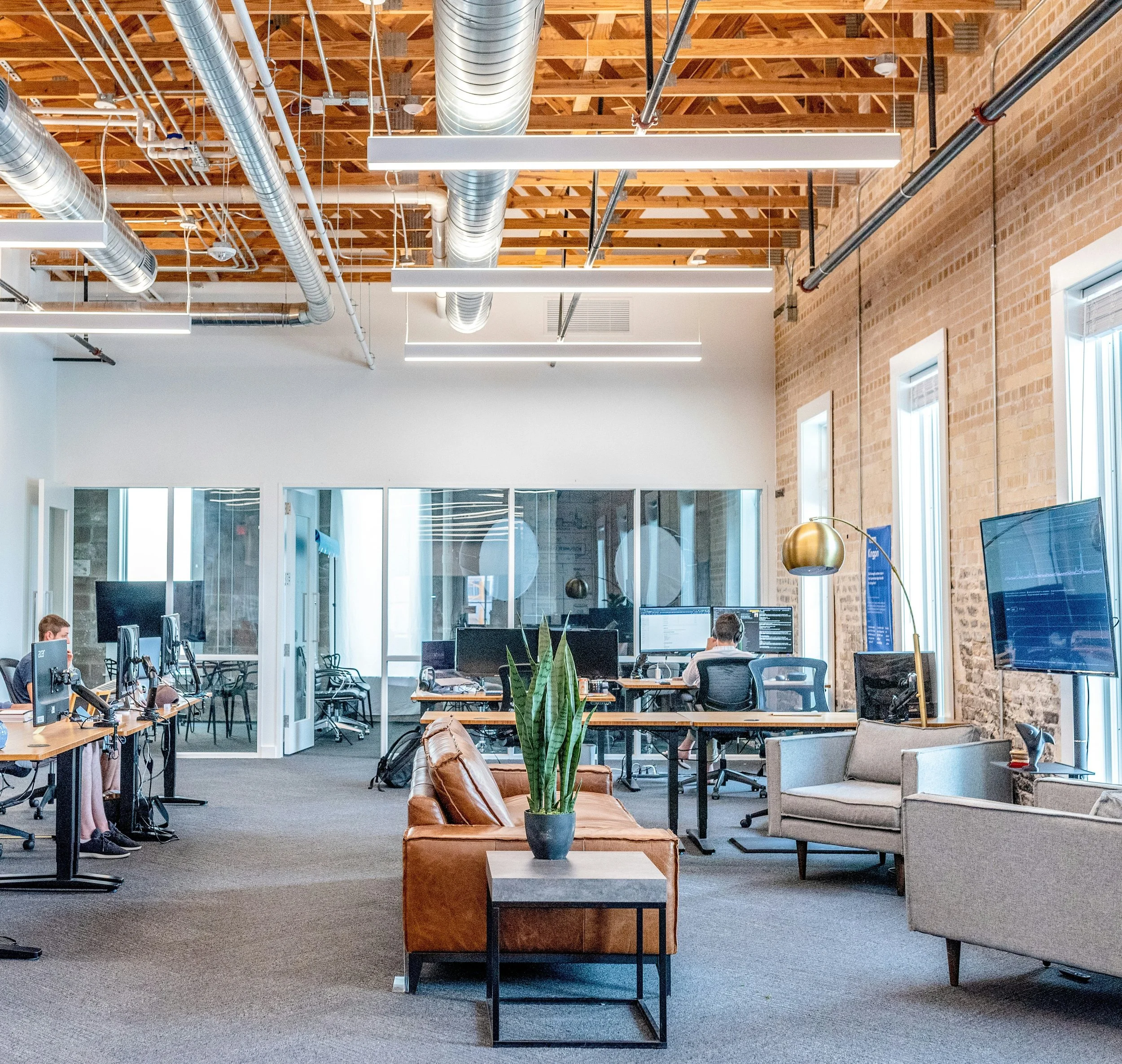 Open-plan office with exposed brick walls, large windows, and modern furniture. Several desks with computers, a leather sofa, a coffee table with a potted plant, and pendant lighting. Employees working at their desks, with visible monitors and office supplies.