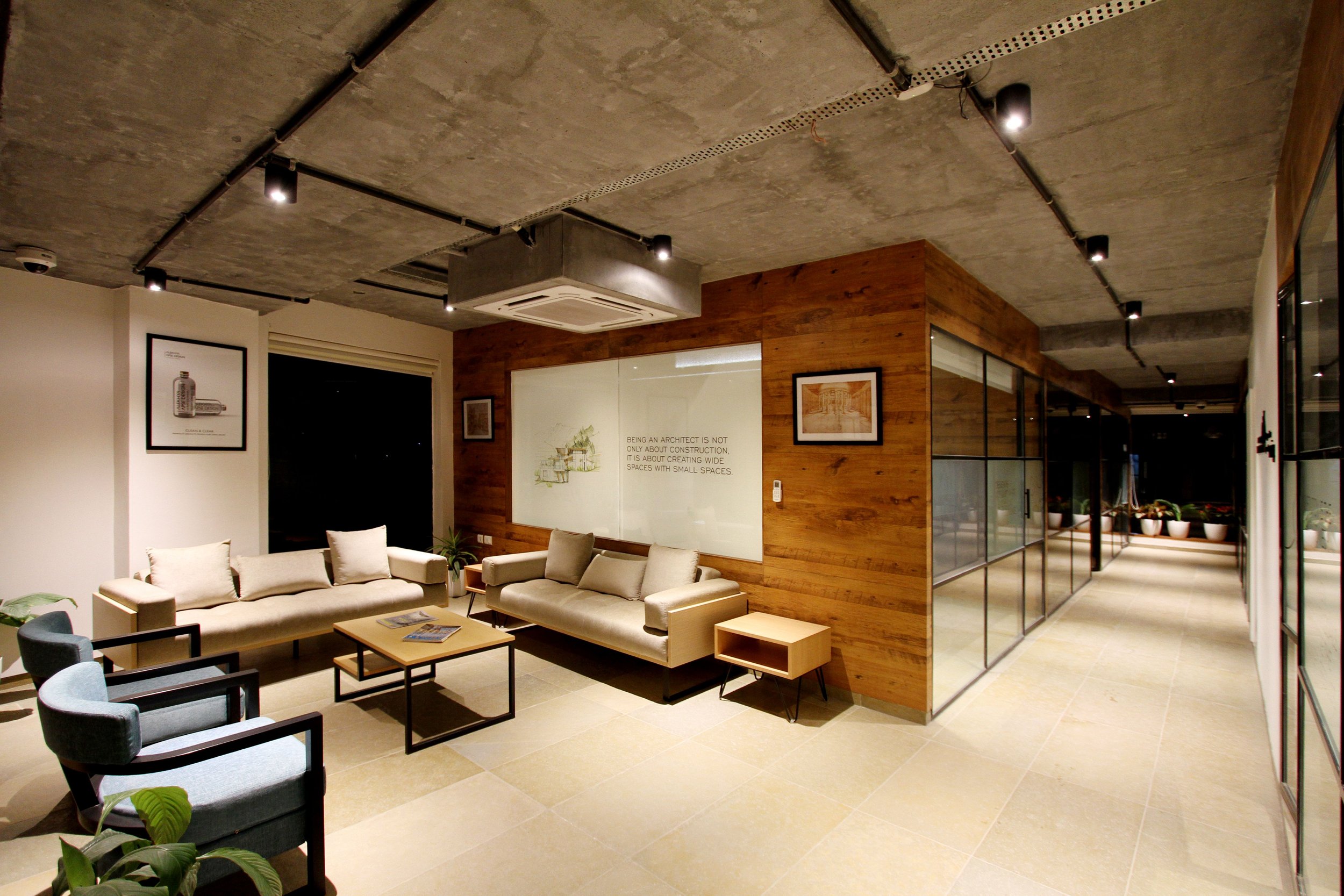 A modern interior room with exposed concrete ceiling, wooden walls, beige tiled floor, and contemporary furnished seating including a beige sectional sofa, chairs, and coffee tables. The room features framed artwork on the walls and potted plants, with a glass partition and framed artwork on wooden walls.