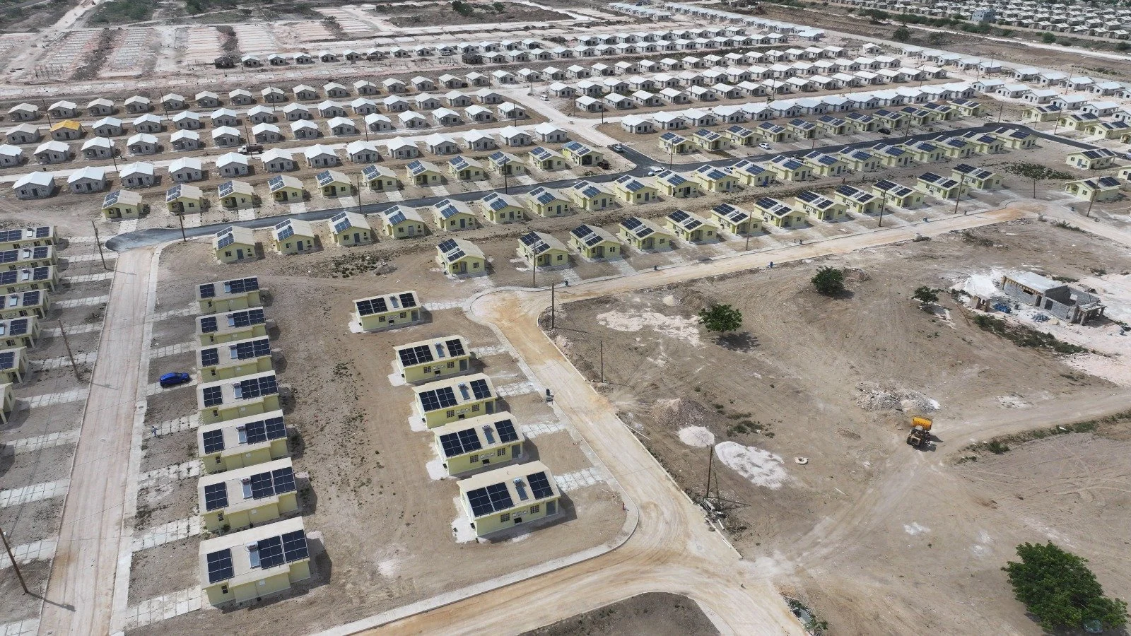 Aerial view of a housing development with many small, similar houses, some with solar panels on the roofs, and some under construction, in a dry, undeveloped area.