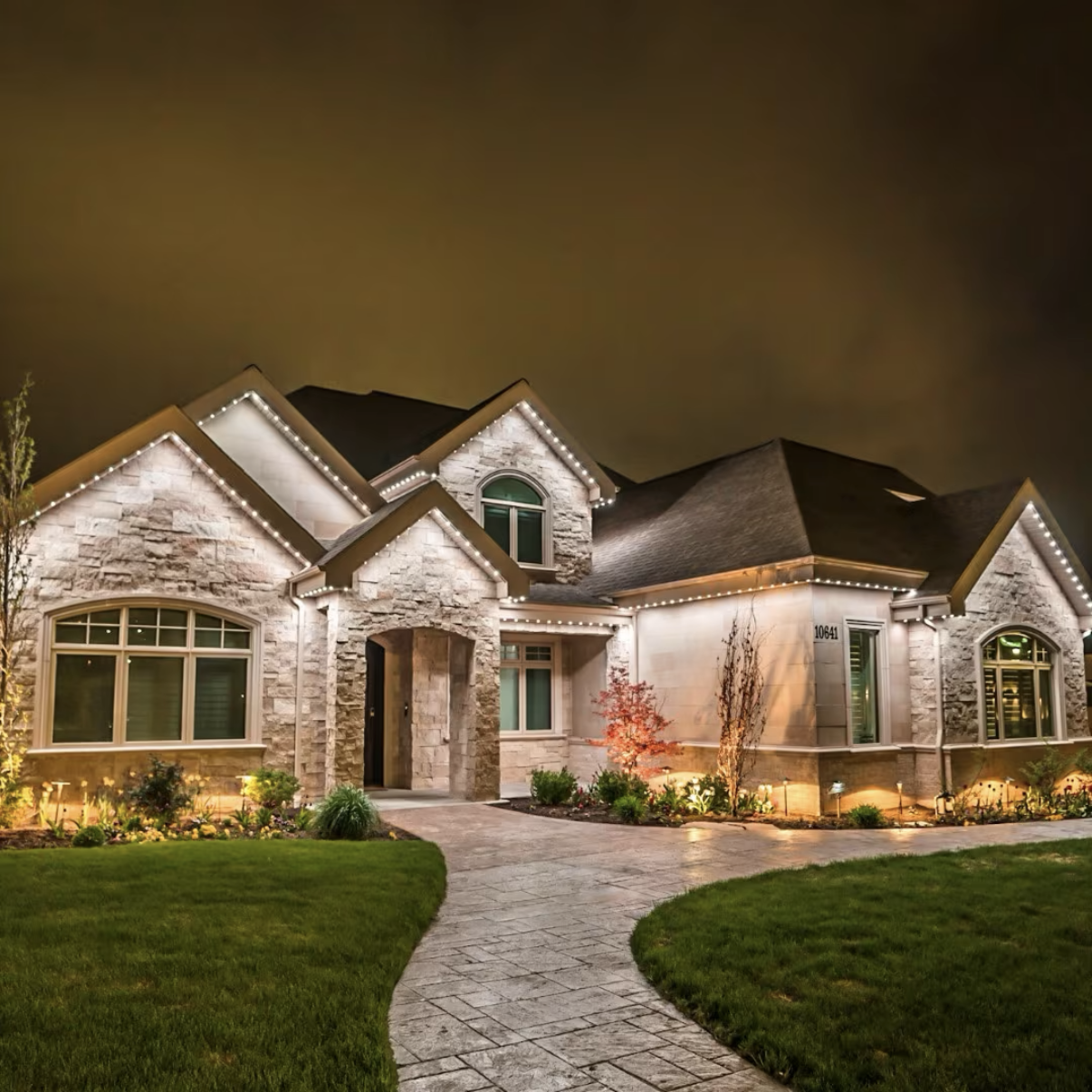 Permanent warm white roofline and landscape lighting on a stone home.