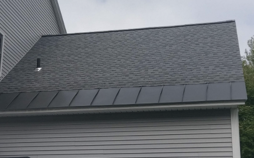Close-up of the roof of a house with asphalt shingles, metal flashing, and a small vent against a partly cloudy sky.