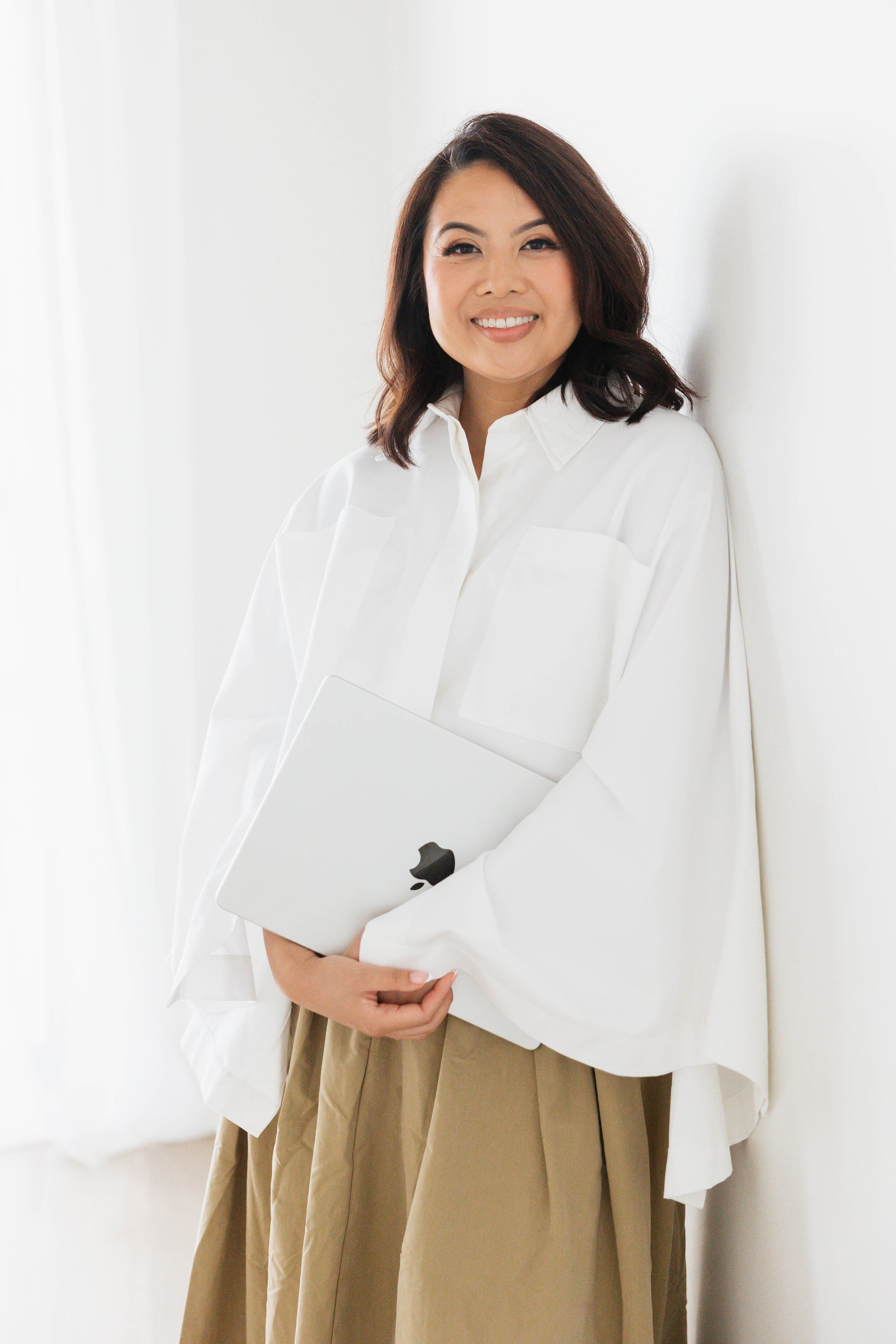 Kristine Allen, hospitality operations consultant and founder of MILA Co Advisory, standing by a white wall. A woman with dark hair in a white blouse, holding a closed laptop.