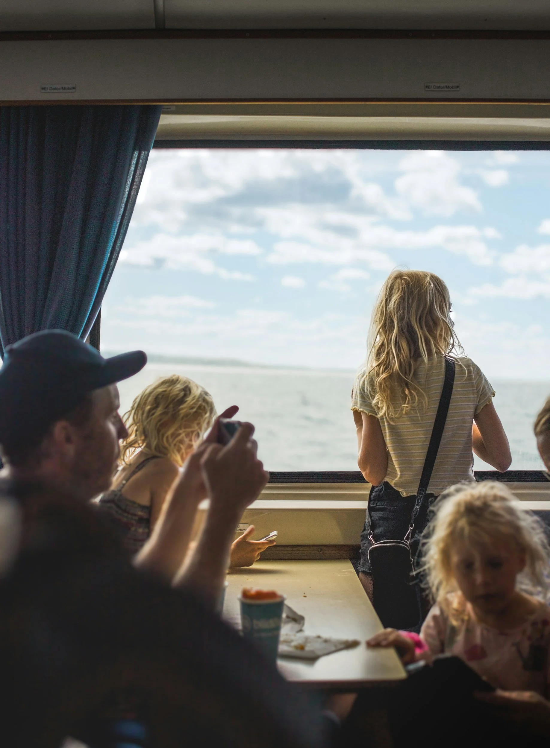 People sitting at a table inside an aircraft, some taking photos or looking outside through the window, with a young woman standing near the window, observing the view of the sky and clouds.