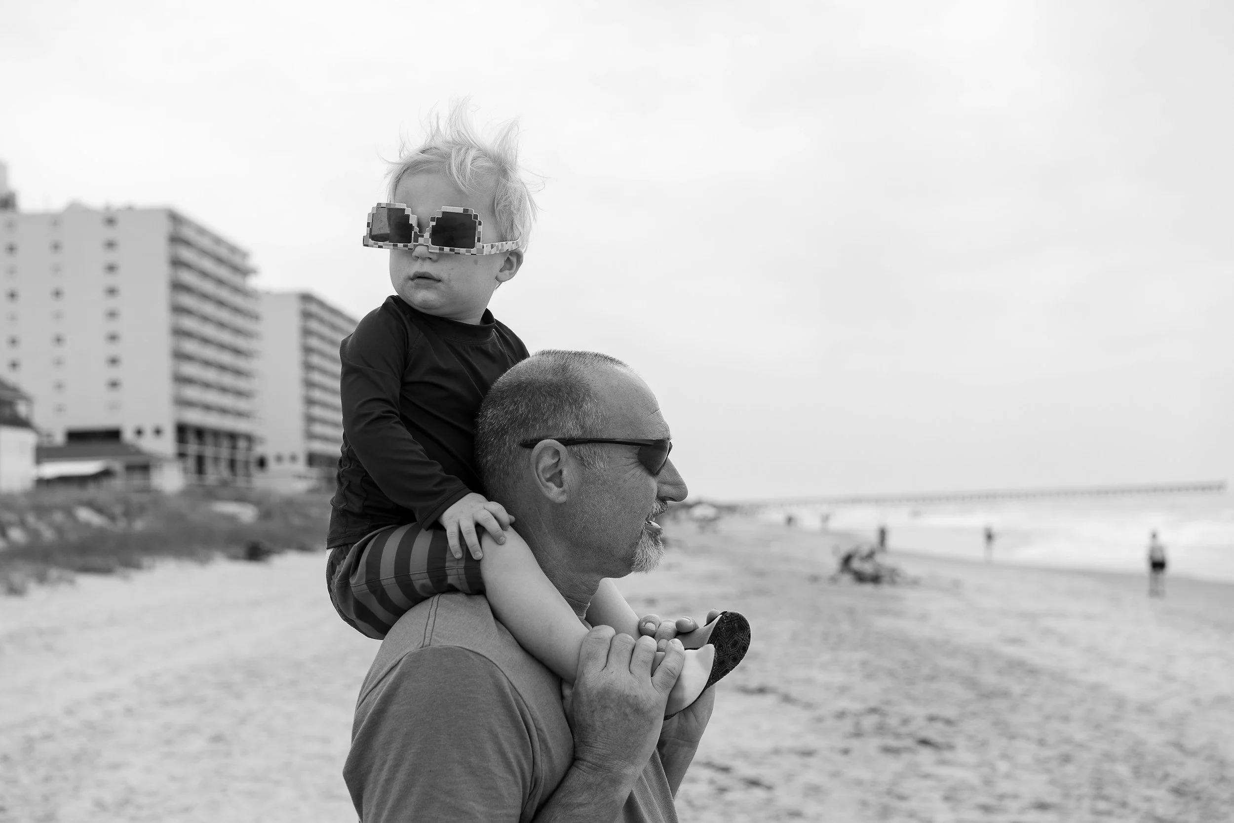 A man carrying a child on his shoulders at the beach, both wearing sunglasses, with buildings and the ocean in the background.