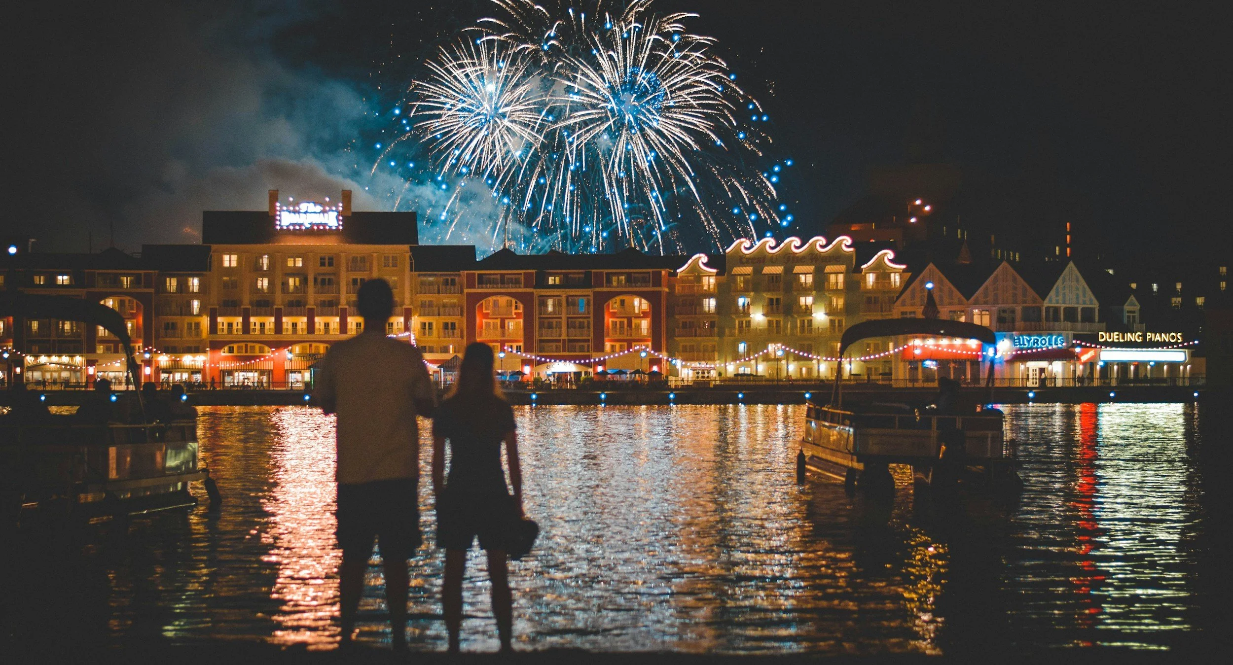 Couple standing by the water watching fireworks display lighting up the night sky over a waterfront cityscape, with colorful lights on buildings and boats reflected in the water.