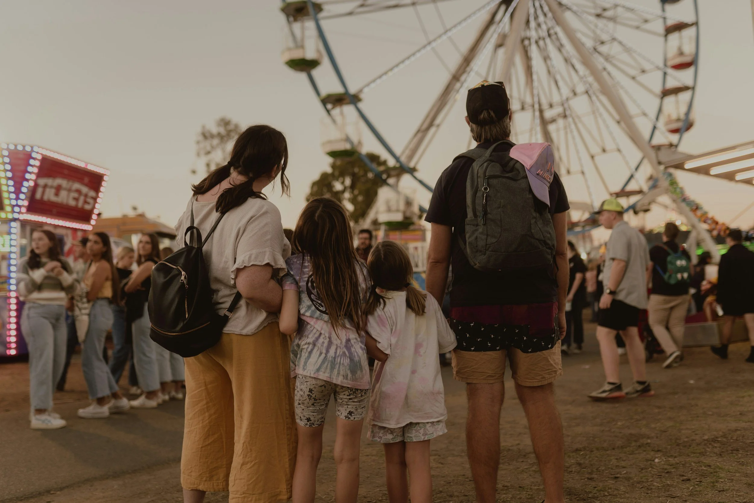 Group of children and adults standing in line at a fairground, with a large Ferris wheel in the background during dusk.