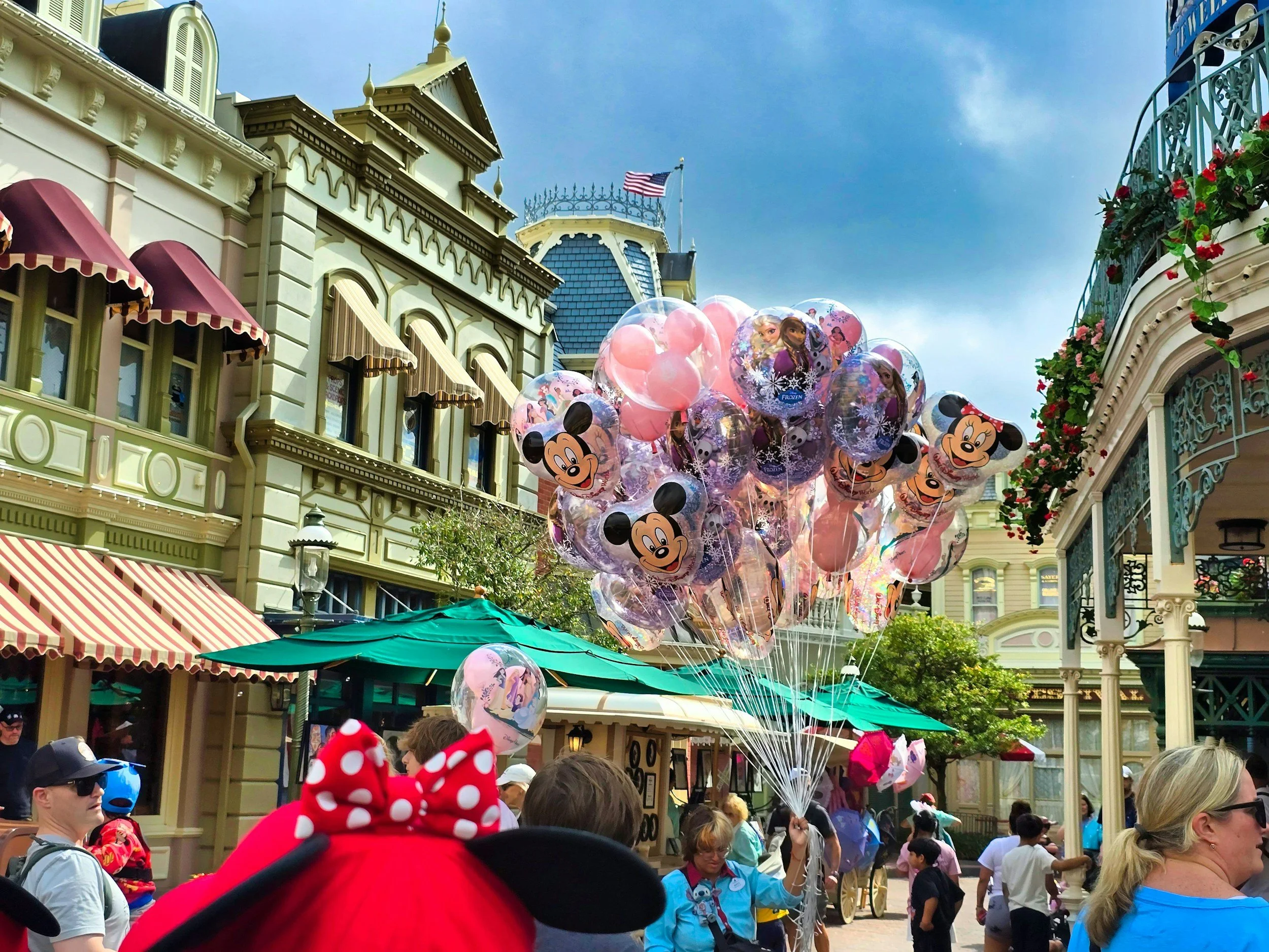 People in Mickey Mouse and Minnie Mouse themed costumes and balloons featuring Disney characters, gathered outside a themed building with Victorian architecture in a Disney park.