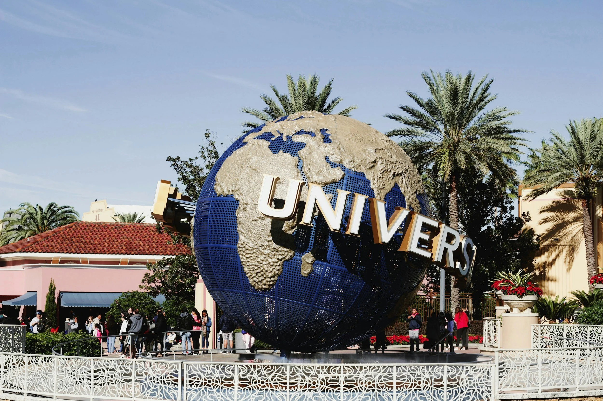 The iconic globe sculpture at Universal Studios theme park with the word 'UNIVERSAL' on it, surrounded by palm trees and people walking around.