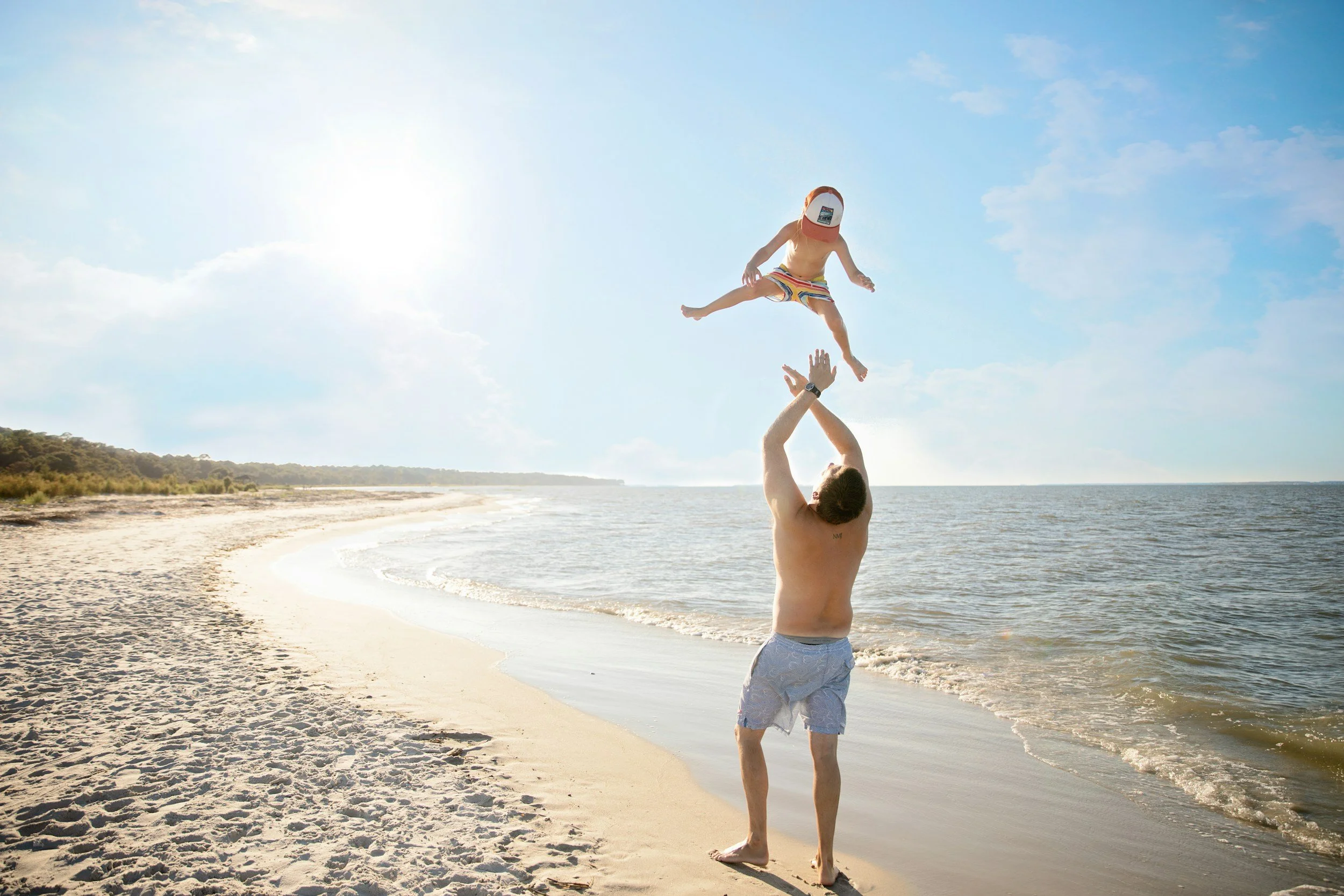 A man lifting a child in the air on a beach with sand and water, under a partly cloudy sky.