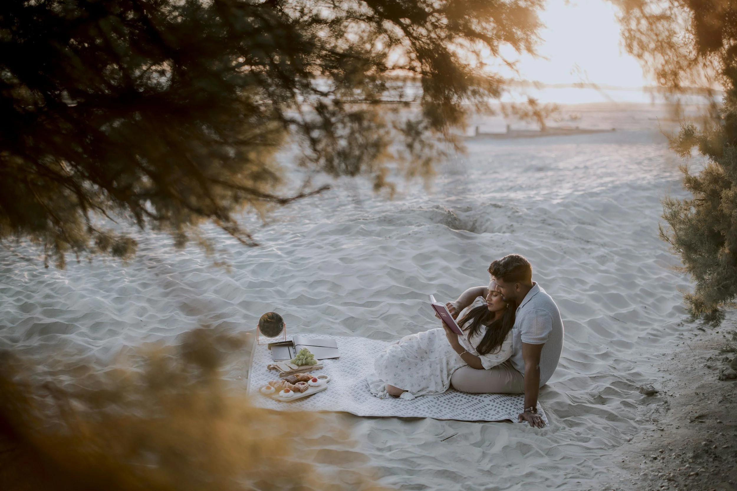 A couple sits on a blanket on a sandy beach, looking at each other affectionately, surrounded by a basket of pastries and snacks.