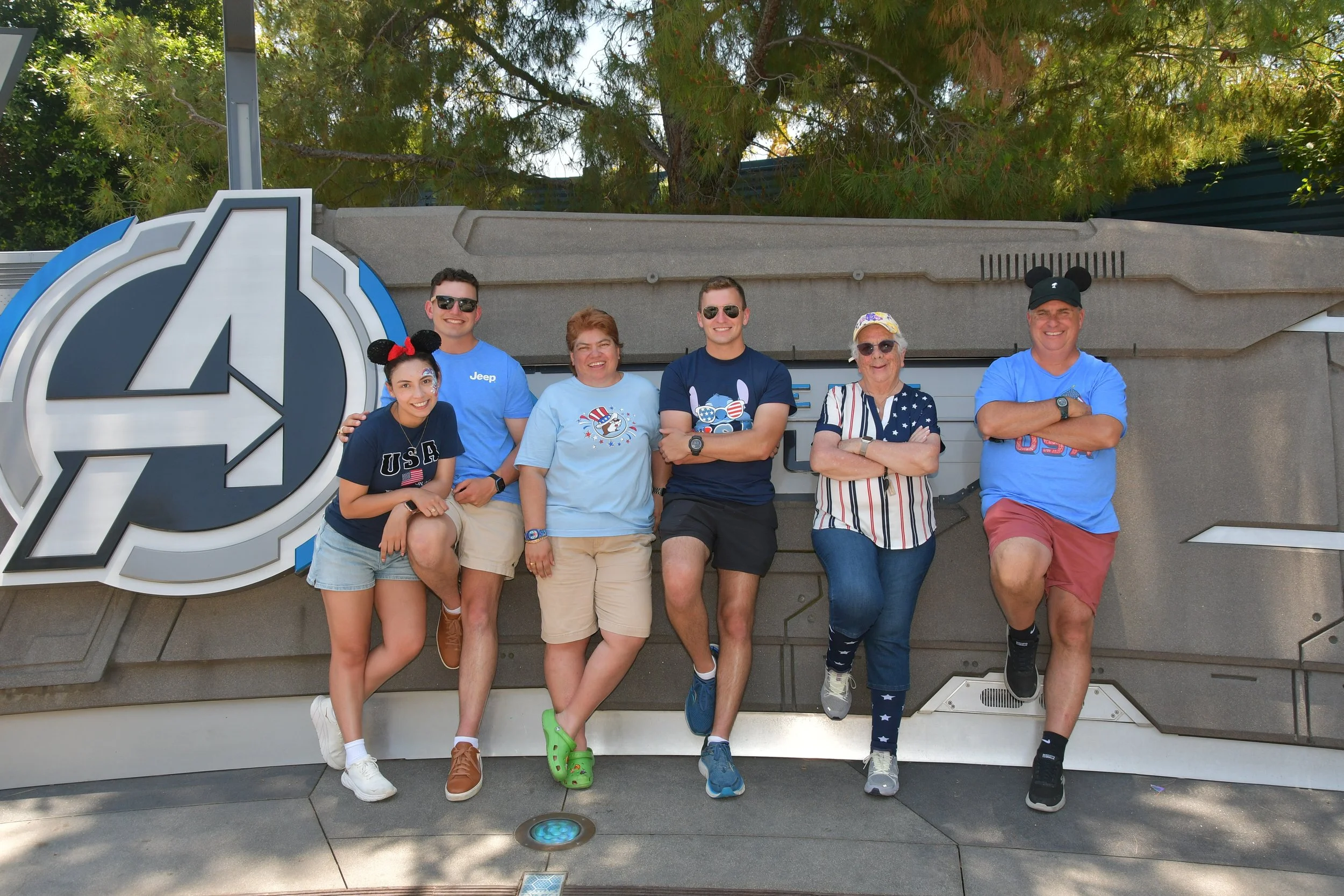 Six people standing in front of a sign at a Disney theme park, some wearing Mickey Mouse ear hats and themed clothing, smiling and posing.