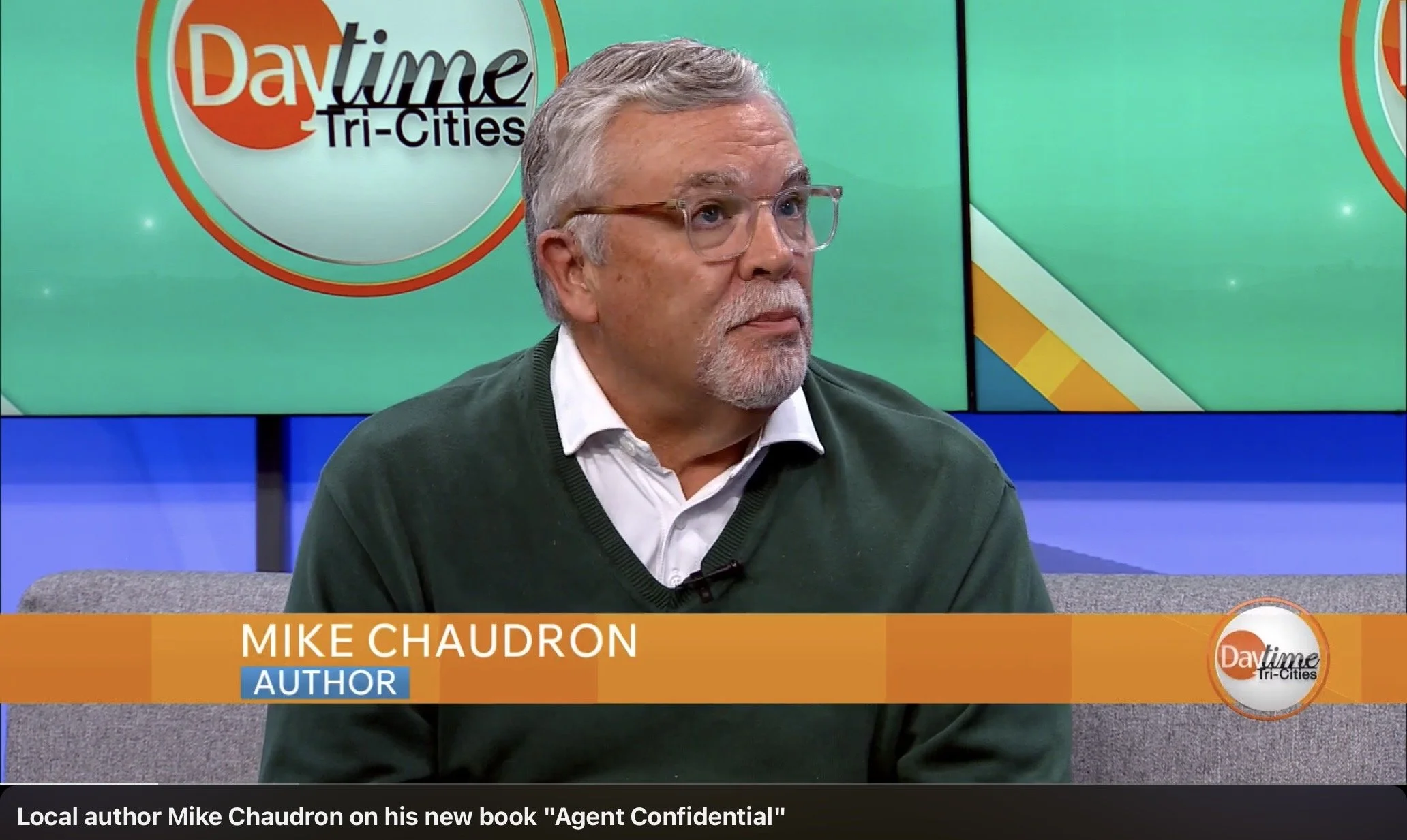 A man with gray hair, beard, and glasses sitting on a television set during an interview. Behind him, there are large screen displays with the text "Daytime Tri-Cities". The lower third graphic shows his name "Mike Chaudron" with the title "Author" and mentions his new book "Agent Confidential".