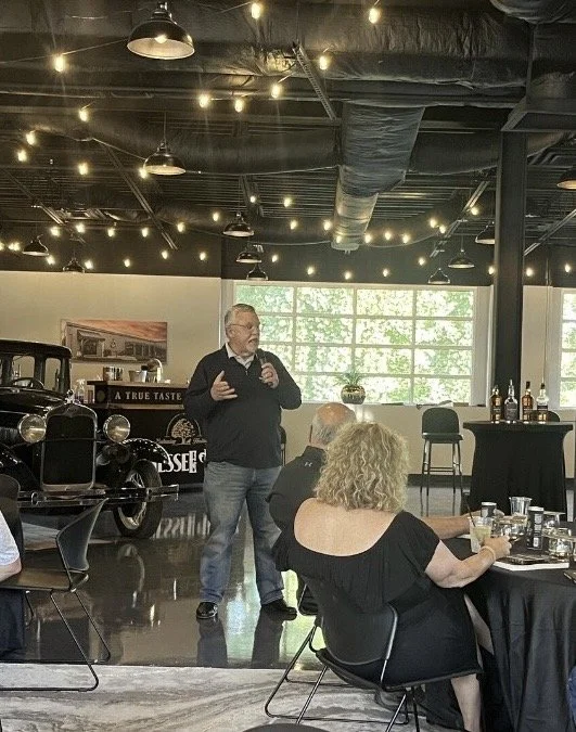 Man giving a presentation to a group in a modern indoor space with vintage car on display, string lights, and large windows.