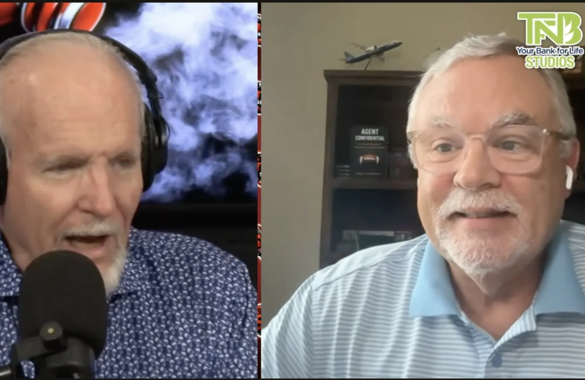 Two middle-aged men with gray hair and glasses engaged in a video call. The man on the left is speaking into a microphone, wearing a dark patterned shirt with a background featuring vape smoke and a red logo. The man on the right is smiling, dressed in a light blue striped shirt, with a bookshelf and model airplane in the background, and a logo reading 'Your Bank for Life Studios' in the top right corner.