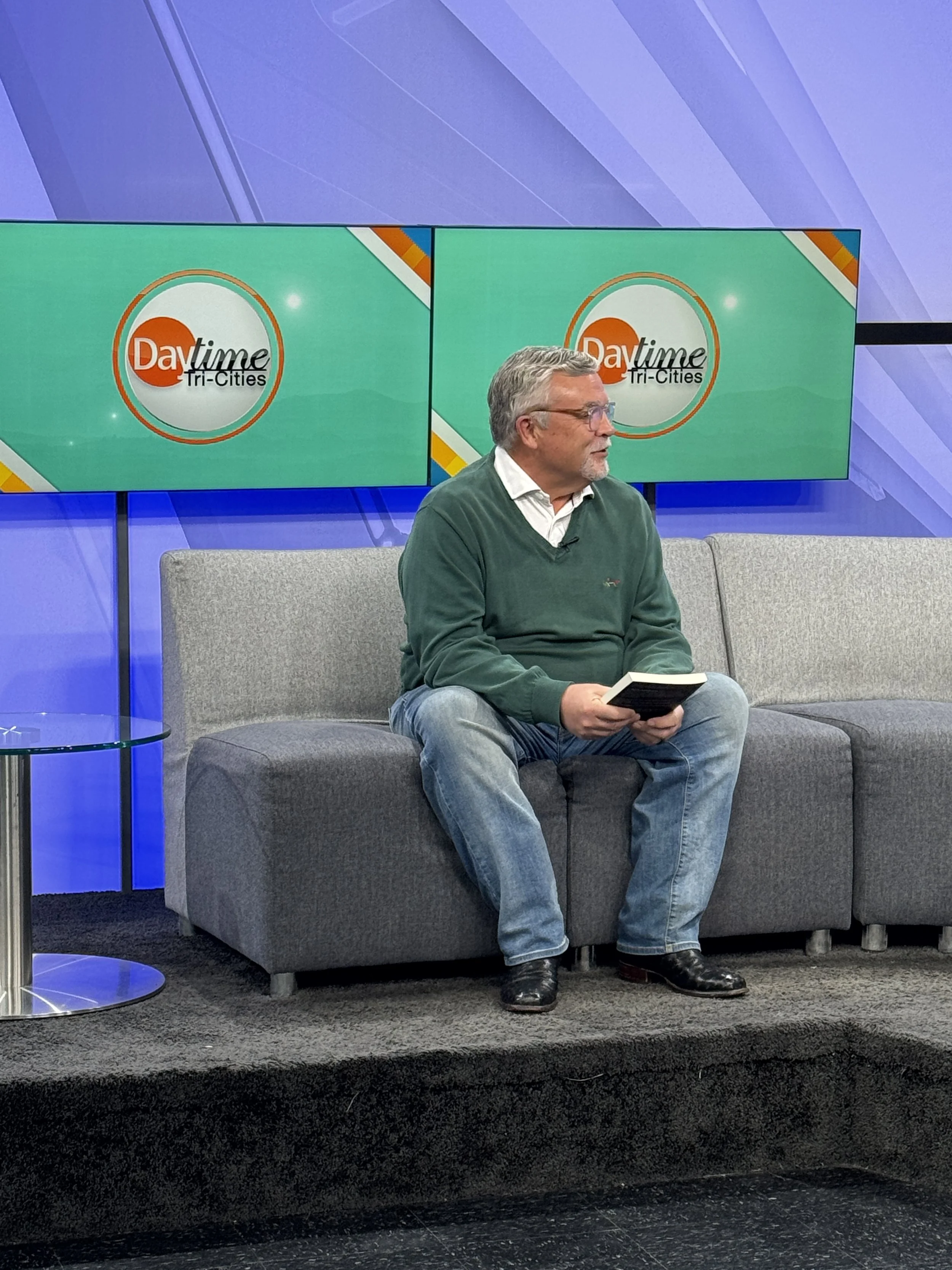 Man sitting on a gray sofa holding a book during a TV interview with a backdrop displaying the 'Daytime Tri-Cities' show logo on two screens.