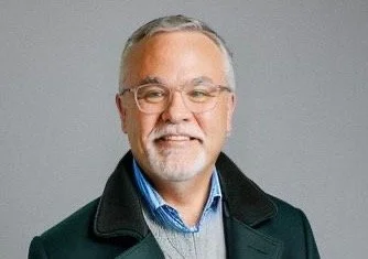Portrait of a smiling older man with glasses, grey hair, and a beard, wearing a dark jacket over a blue shirt, against a grey background.