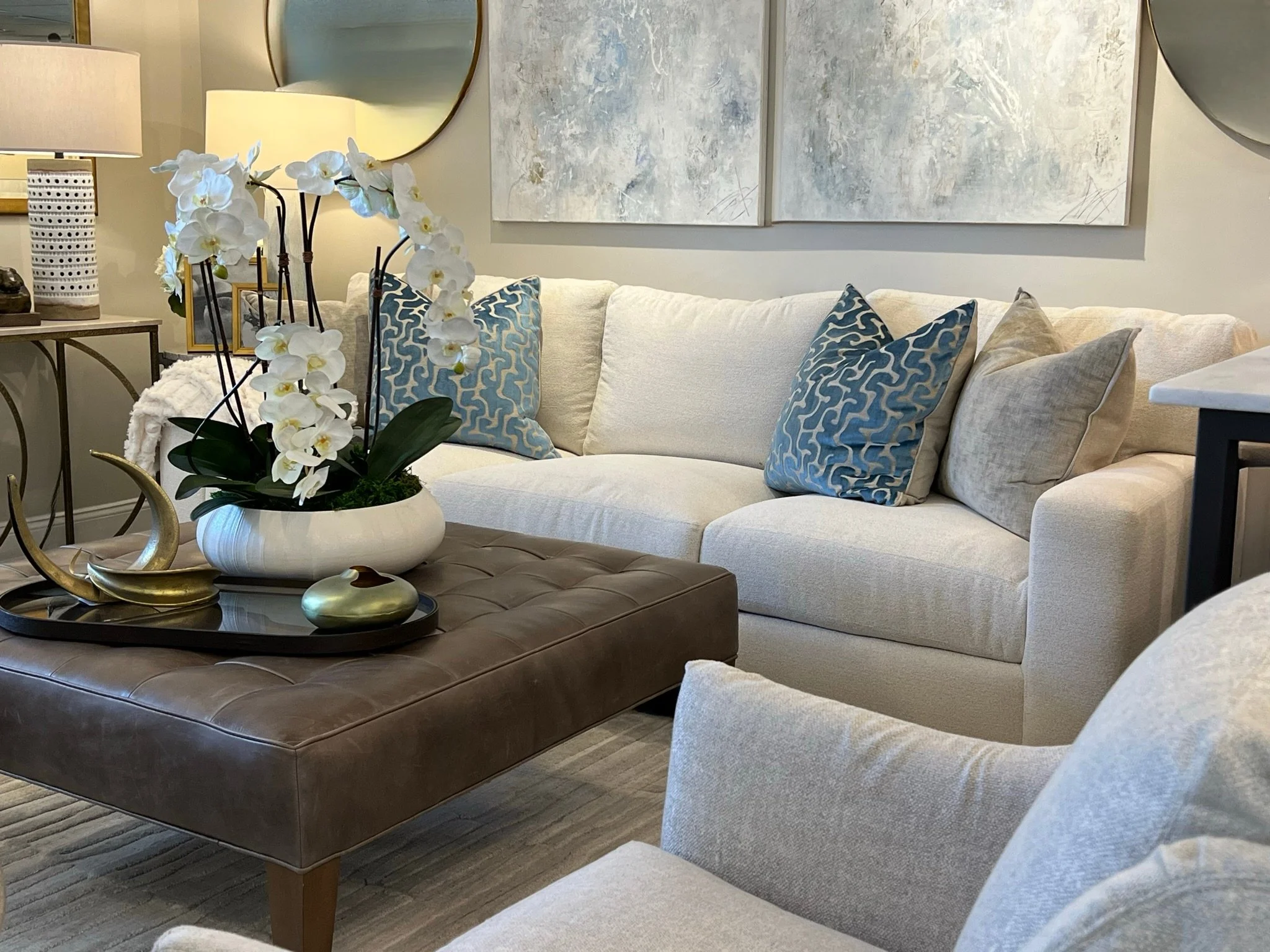 Living room with white sectional sofa, blue patterned pillows, and a round coffee table with a white orchid arrangement. There is a side table with a lamp and decorative items, and abstract artwork on the wall.